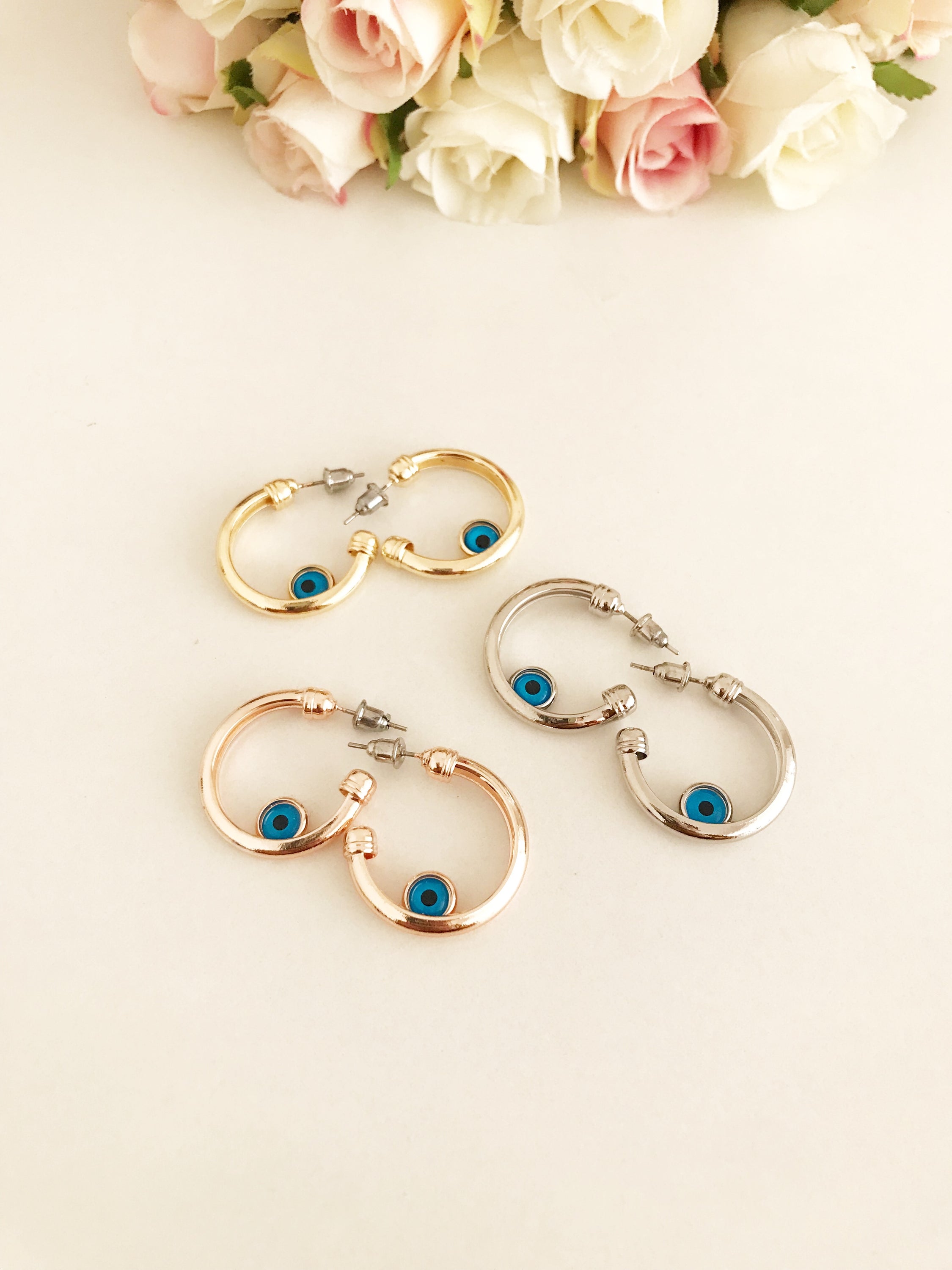 A pair of handmade evil eye hoop earrings featuring a blue evil eye charm, available in rose gold, gold, and silver finishes.