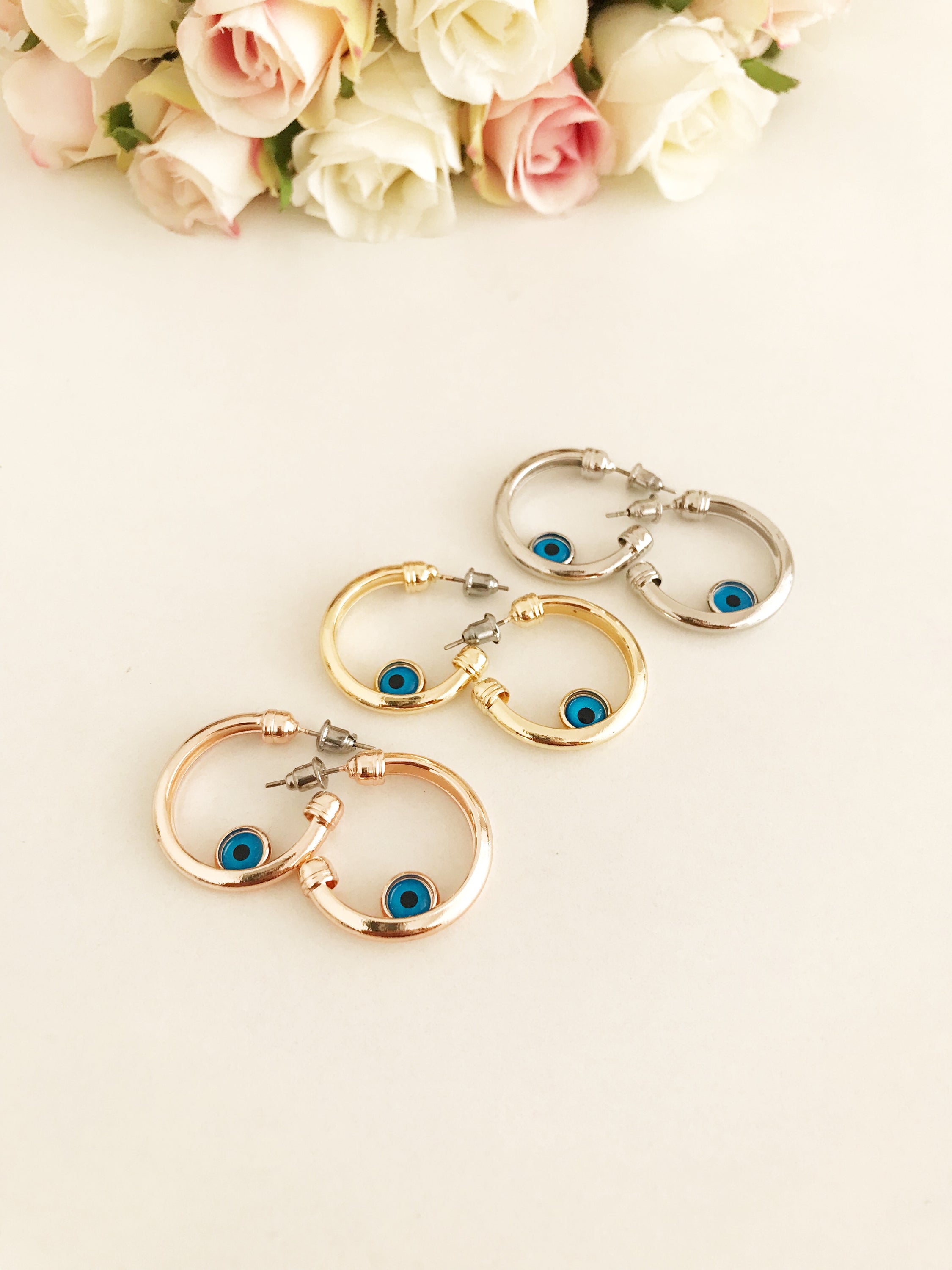 A pair of handmade evil eye hoop earrings featuring a blue evil eye charm, available in rose gold, gold, and silver finishes.