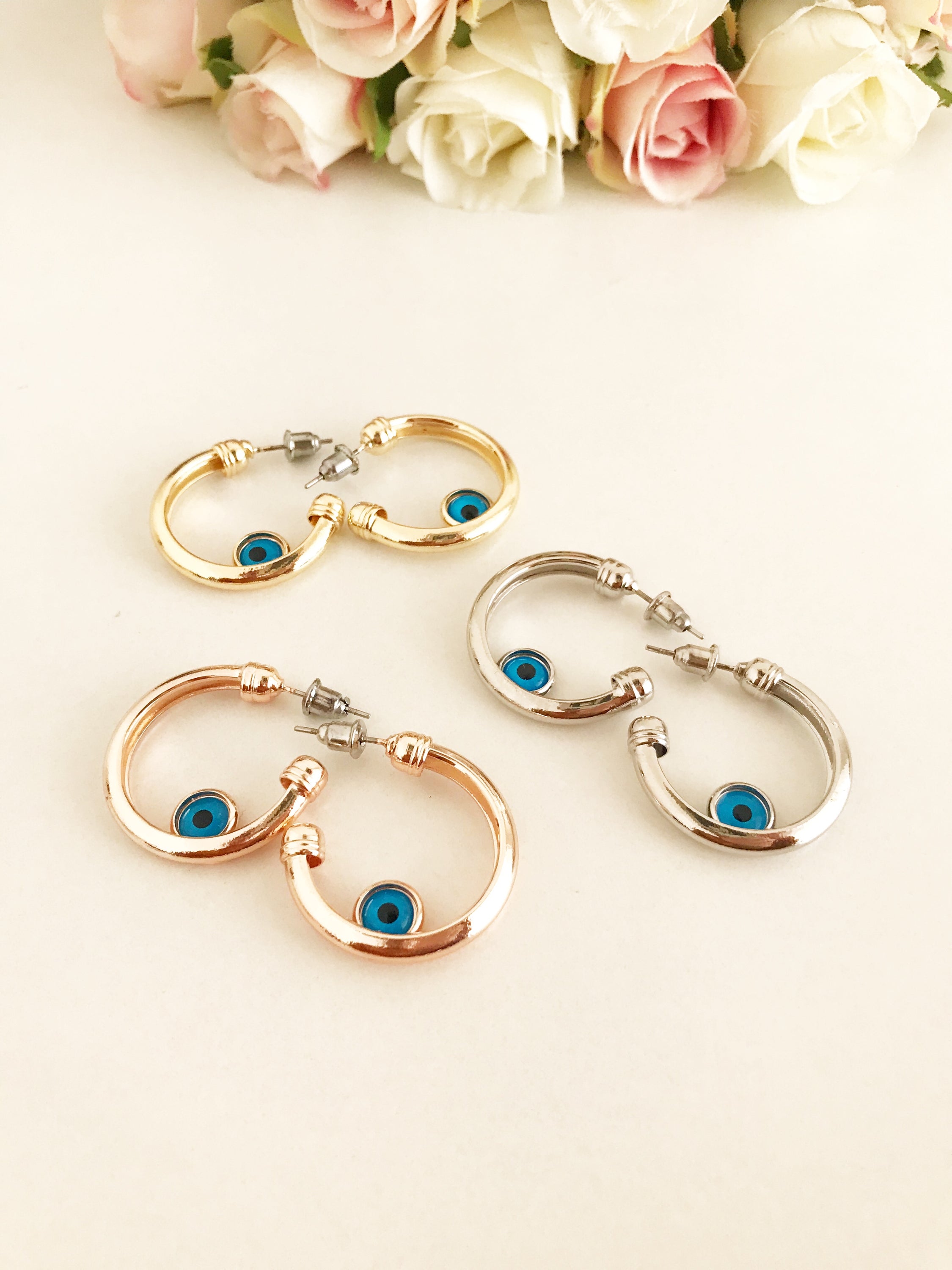 A pair of handmade evil eye hoop earrings featuring a blue evil eye charm, available in rose gold, gold, and silver finishes.