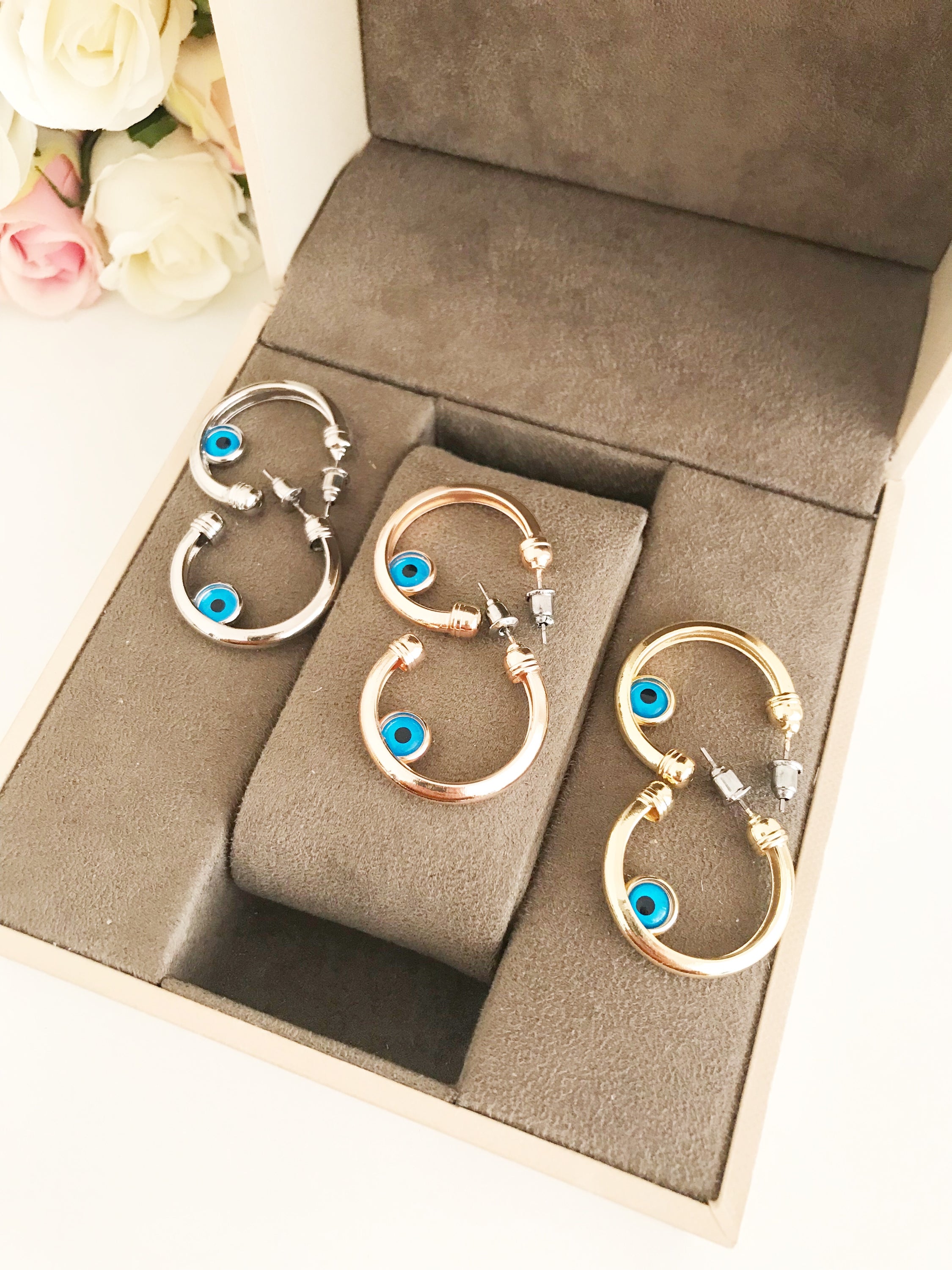 A pair of handmade evil eye hoop earrings featuring a blue evil eye charm, available in rose gold, gold, and silver finishes.