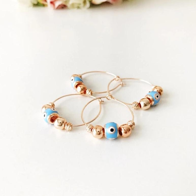 A pair of handmade Evil Eye Turquoise Hoop Earrings featuring a blue evil eye bead and white glass charm, set in a rose gold finish.