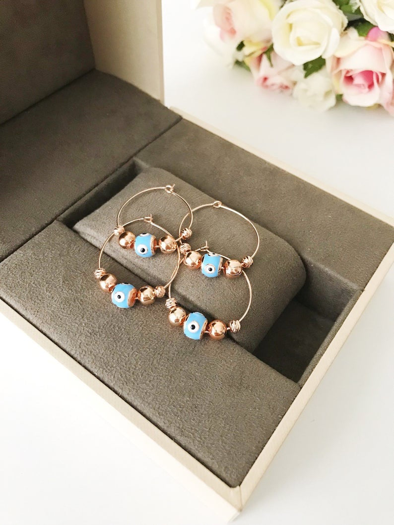 A pair of handmade Evil Eye Turquoise Hoop Earrings featuring a blue evil eye bead and white glass charm, set in a rose gold finish.