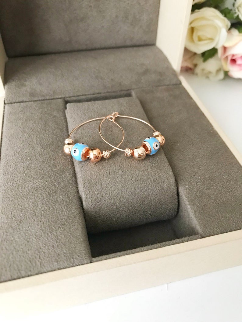 A pair of handmade Evil Eye Turquoise Hoop Earrings featuring a blue evil eye bead and white glass charm, set in a rose gold finish.