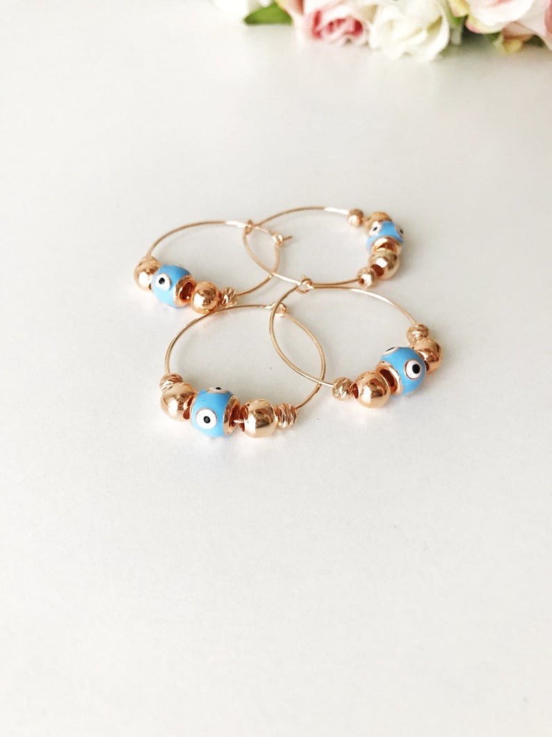 A pair of handmade Evil Eye Turquoise Hoop Earrings featuring a blue evil eye bead and white glass charm, set in a rose gold finish.