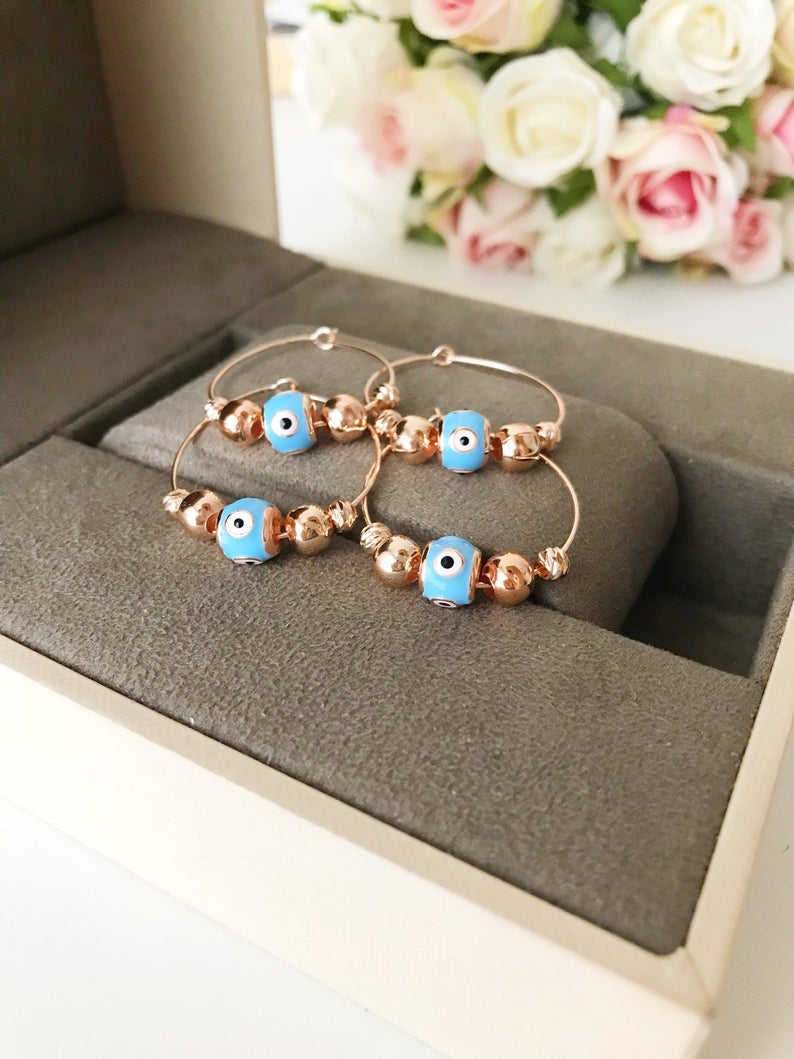 A pair of handmade Evil Eye Turquoise Hoop Earrings featuring a blue evil eye bead and white glass charm, set in a rose gold finish.