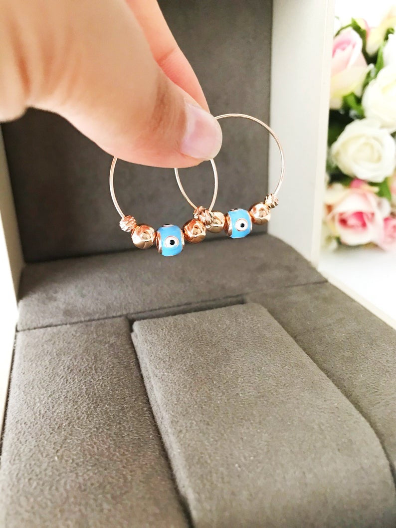 A pair of handmade Evil Eye Turquoise Hoop Earrings featuring a blue evil eye bead and white glass charm, set in a rose gold finish.