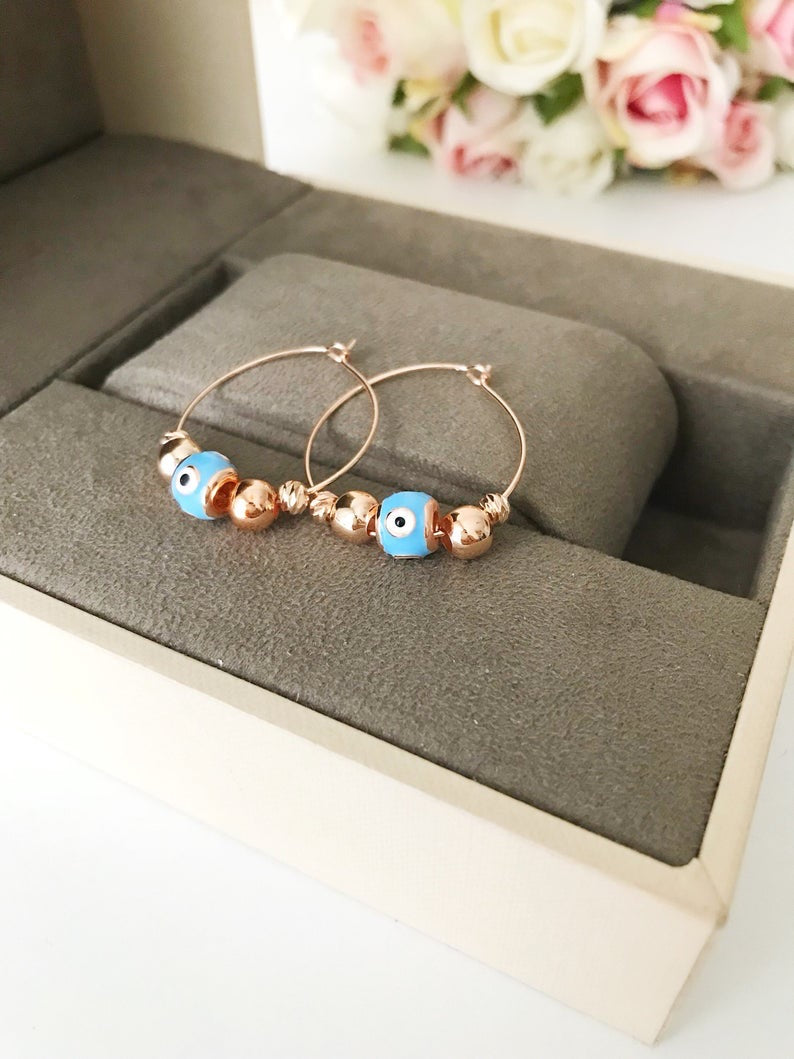 A pair of handmade Evil Eye Turquoise Hoop Earrings featuring a blue evil eye bead and white glass charm, set in a rose gold finish.