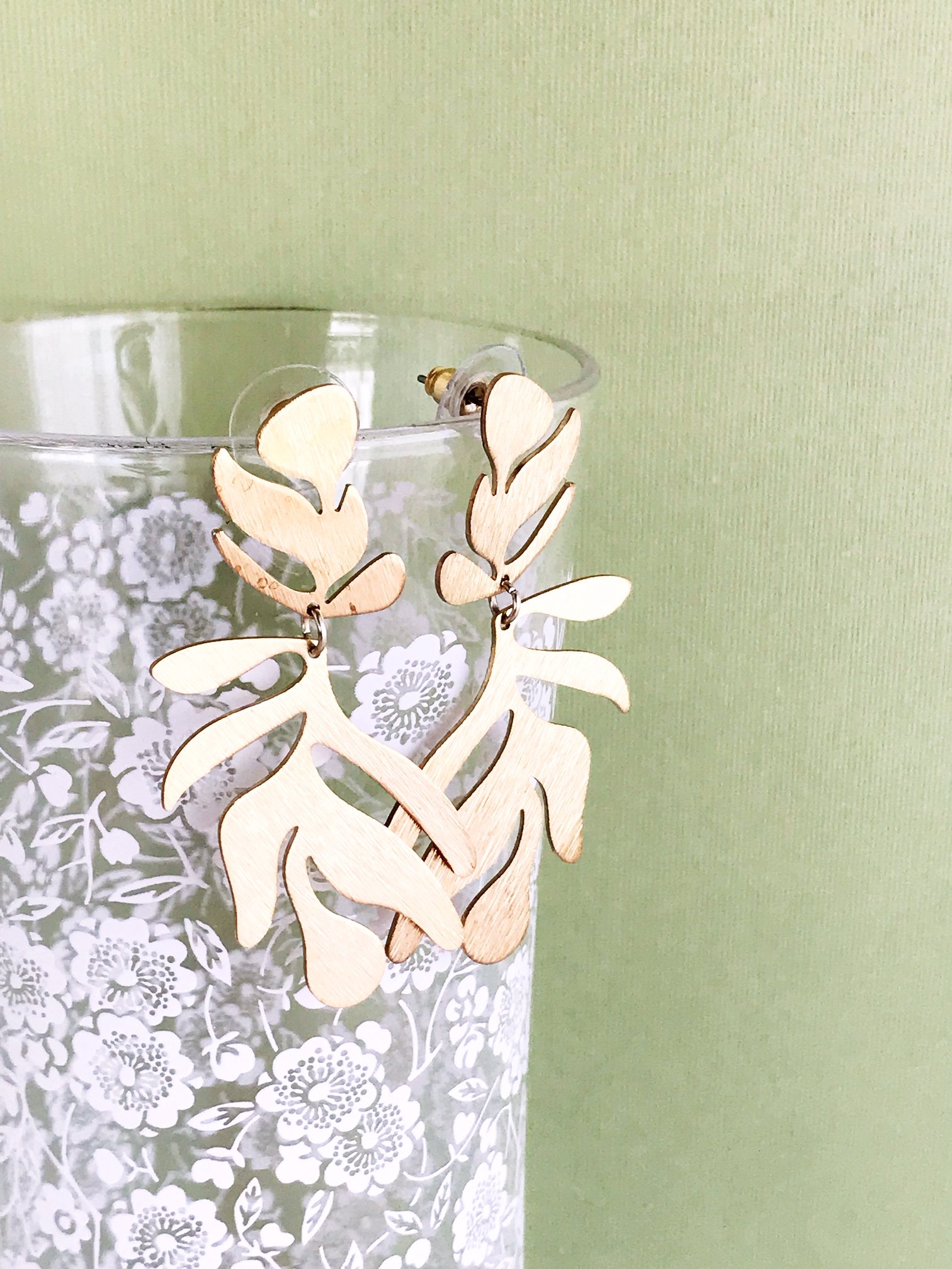 A pair of lightweight floral-inspired frond earrings in brushed brass, showcasing their elegant design and stainless steel ear posts.