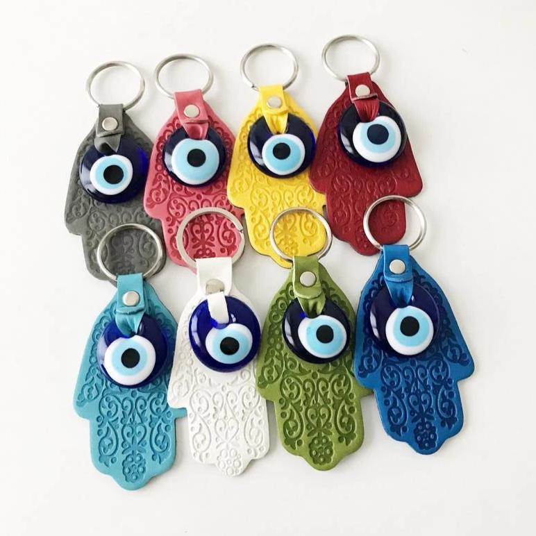 A stylish Hamsa hand evil eye keychain made of faux leather with a blue glass evil eye bead, available in various colors.