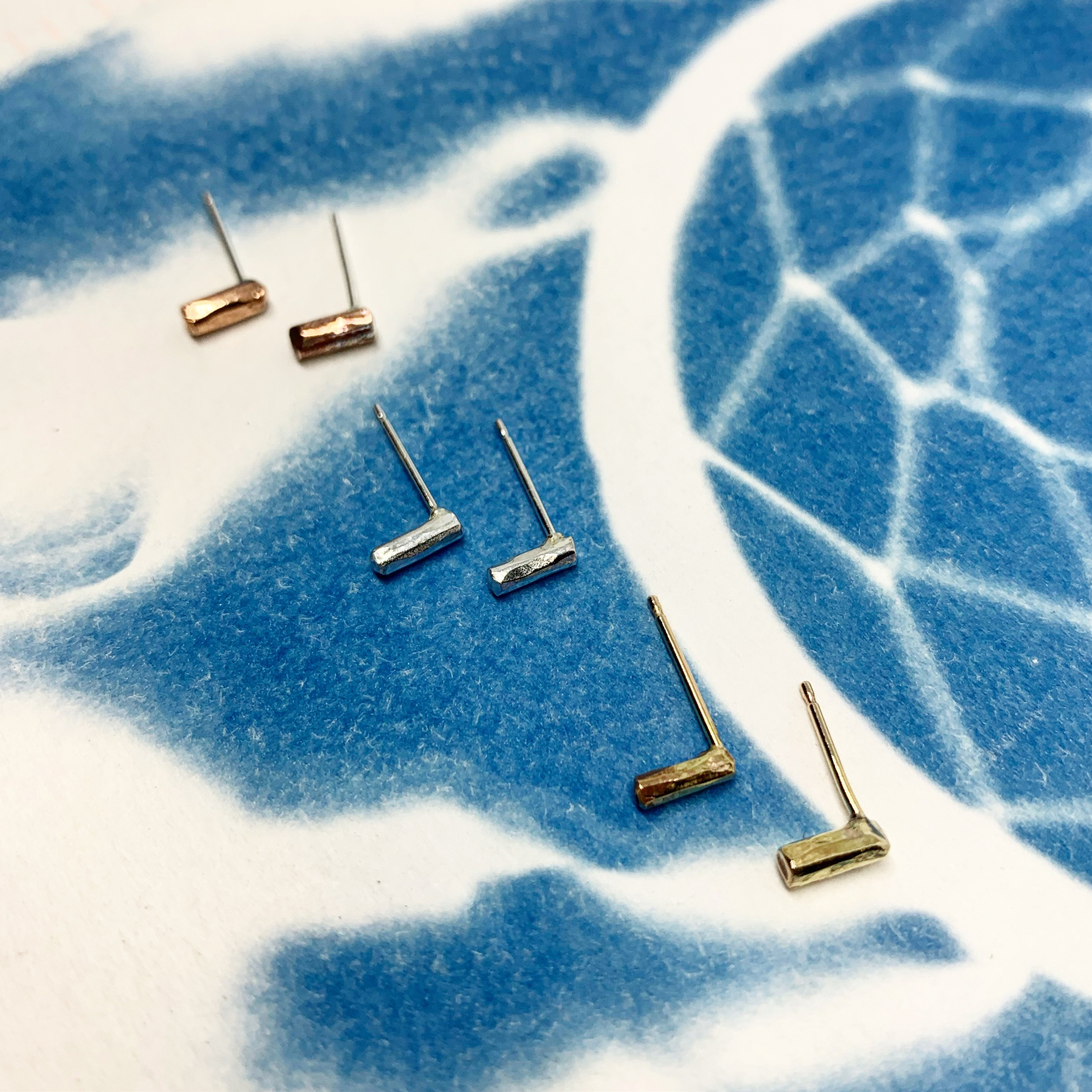 A pair of Mini Balance Stud Earrings - 1/4" featuring tiny hammered rods in copper, sterling silver, and yellow gold fill options, showcasing their minimalist design.