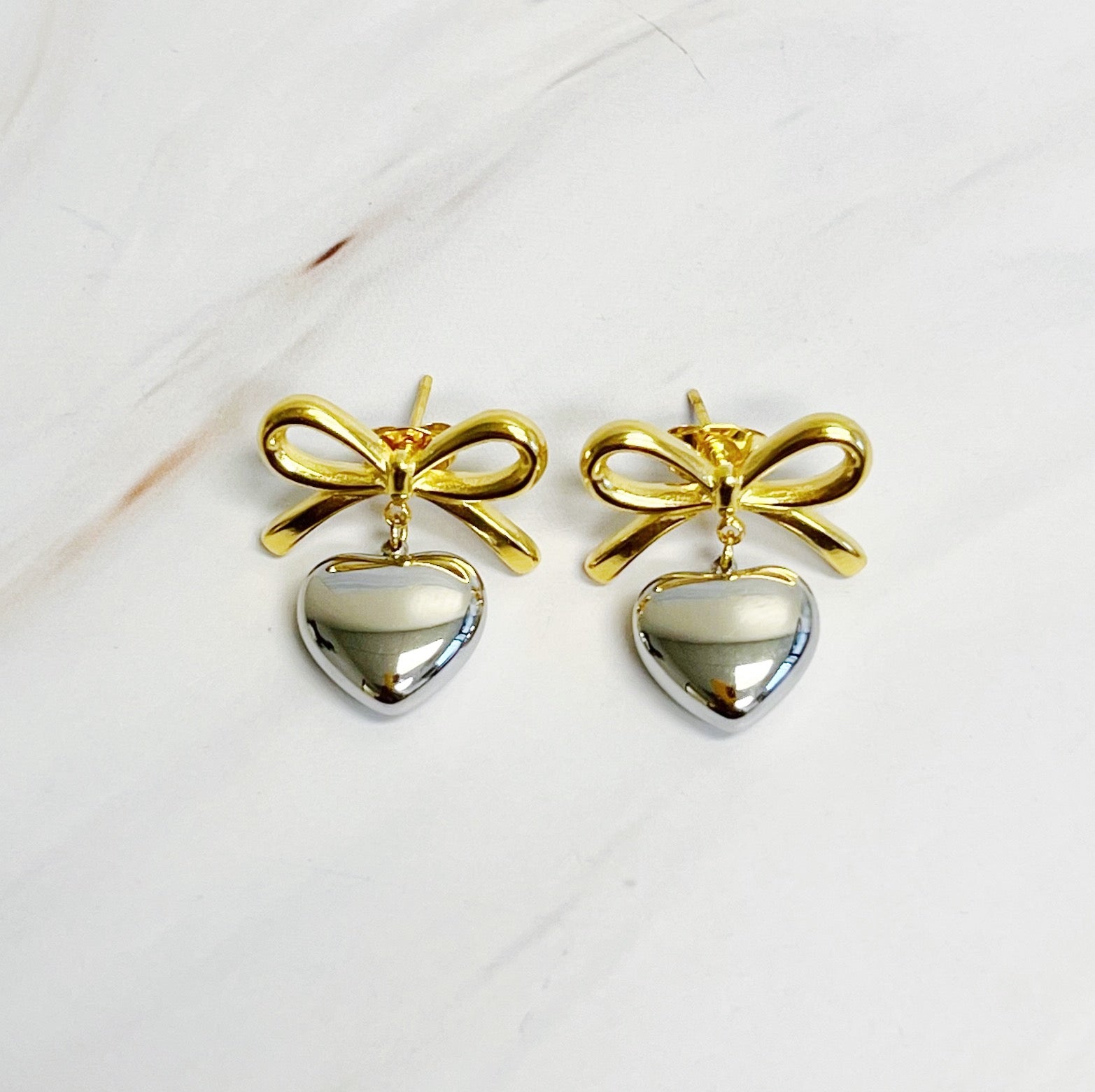 A pair of Mini Bow Mini Heart Earrings featuring a delicate bow and heart design, crafted from 18k gold plated stainless steel.