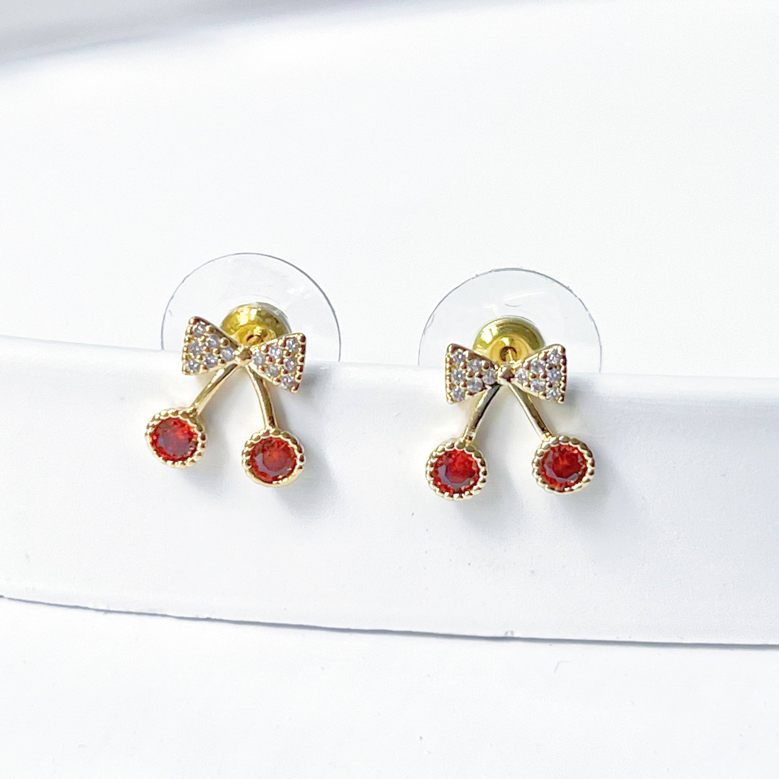 A pair of Mini Red Cherry Stud Earrings featuring red zircon cherries and clear zircon bow ties, set on a gold-plated bronze base with sterling silver studs.