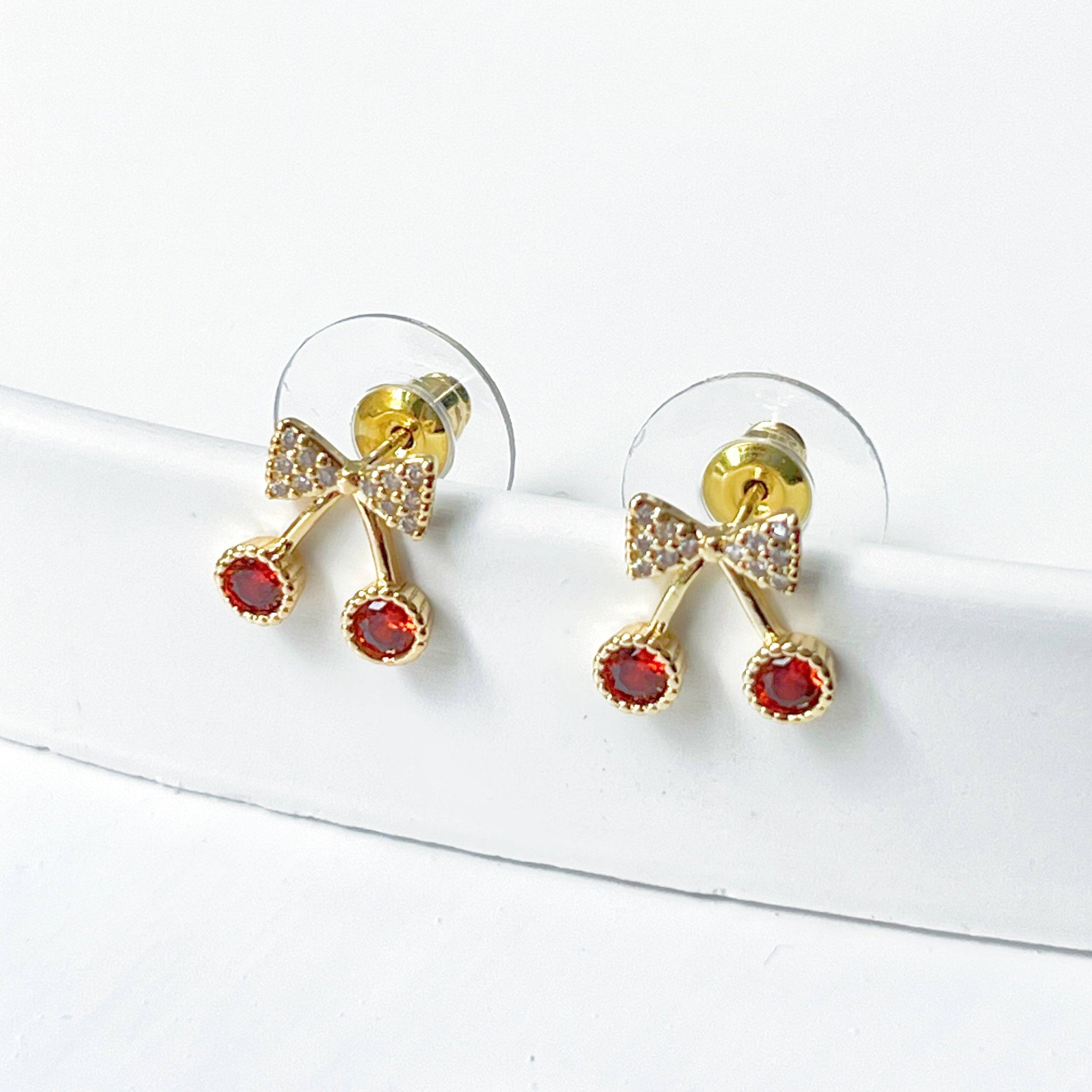 A pair of Mini Red Cherry Stud Earrings featuring red zircon cherries and clear zircon bow ties, set on a gold-plated bronze base with sterling silver studs.