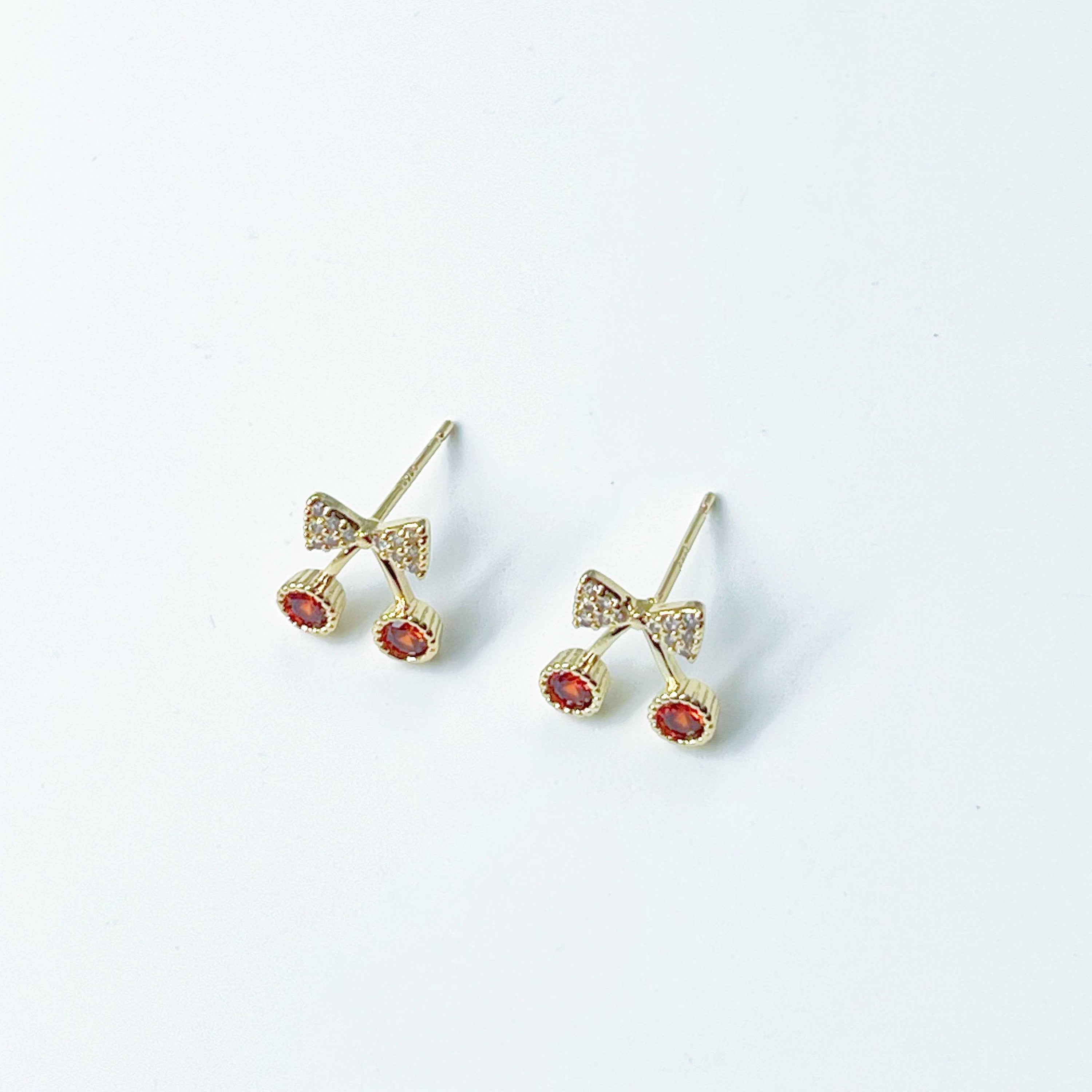 A pair of Mini Red Cherry Stud Earrings featuring red zircon cherries and clear zircon bow ties, set on a gold-plated bronze base with sterling silver studs.