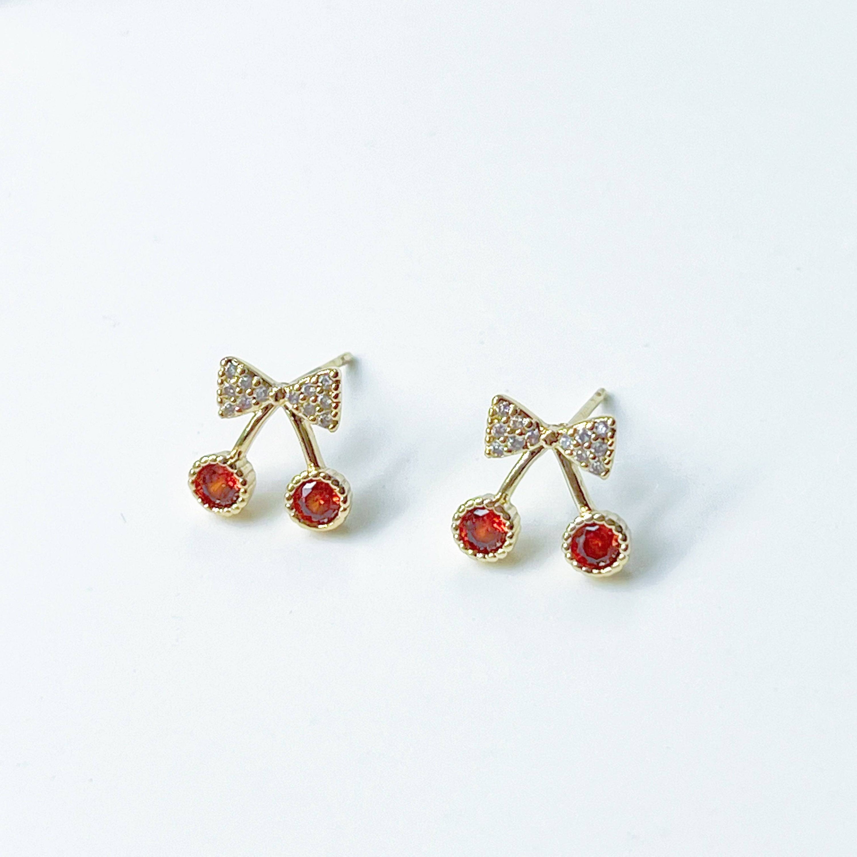 A pair of Mini Red Cherry Stud Earrings featuring red zircon cherries and clear zircon bow ties, set on a gold-plated bronze base with sterling silver studs.