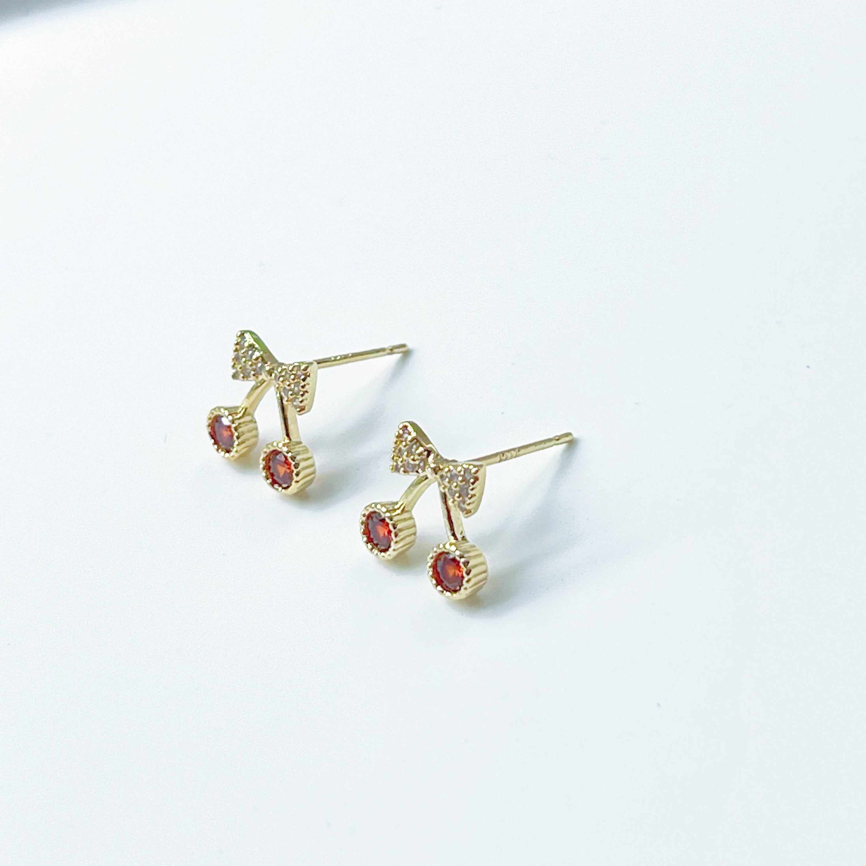 A pair of Mini Red Cherry Stud Earrings featuring red zircon cherries and clear zircon bow ties, set on a gold-plated bronze base with sterling silver studs.