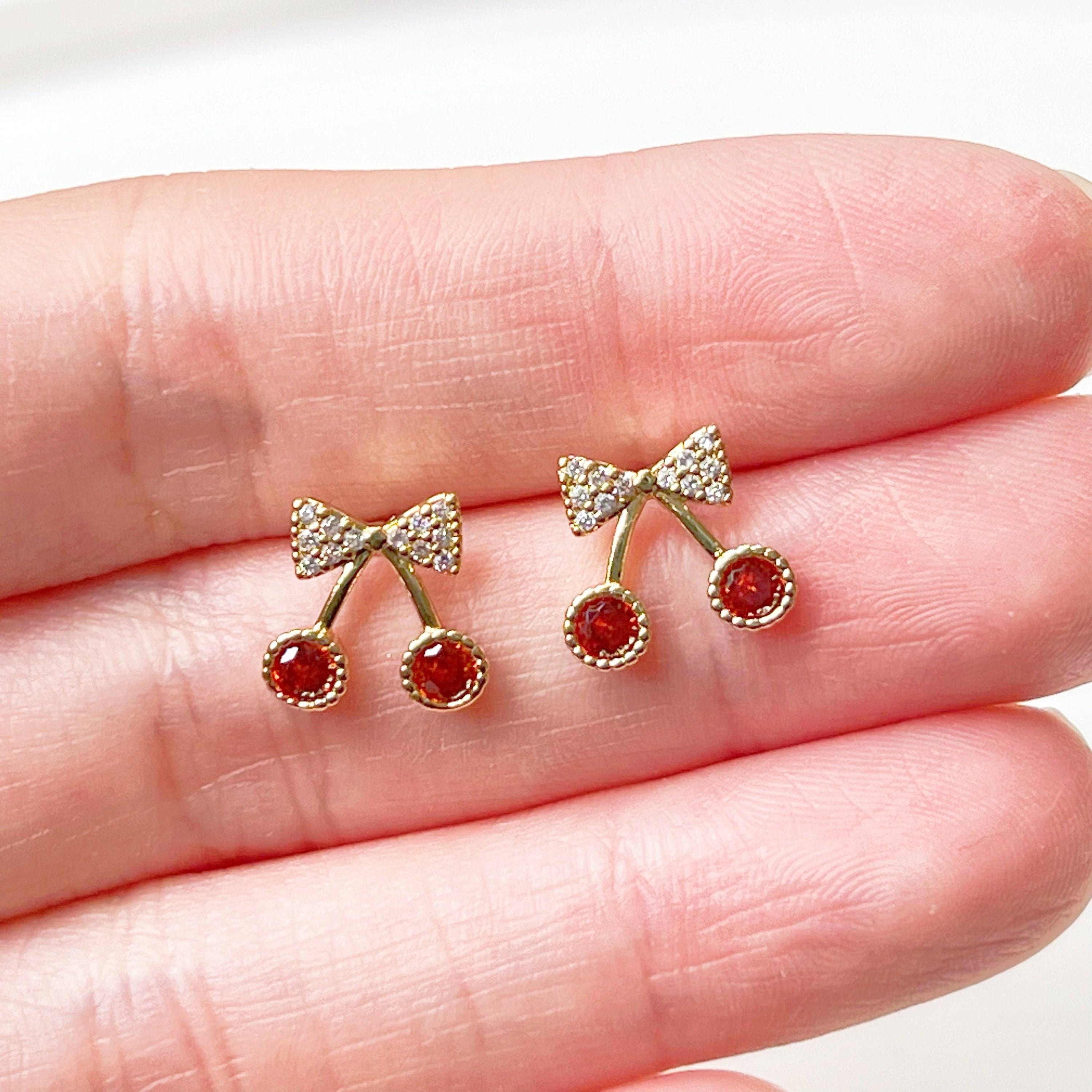 A pair of Mini Red Cherry Stud Earrings featuring red zircon cherries and clear zircon bow ties, set on a gold-plated bronze base with sterling silver studs.