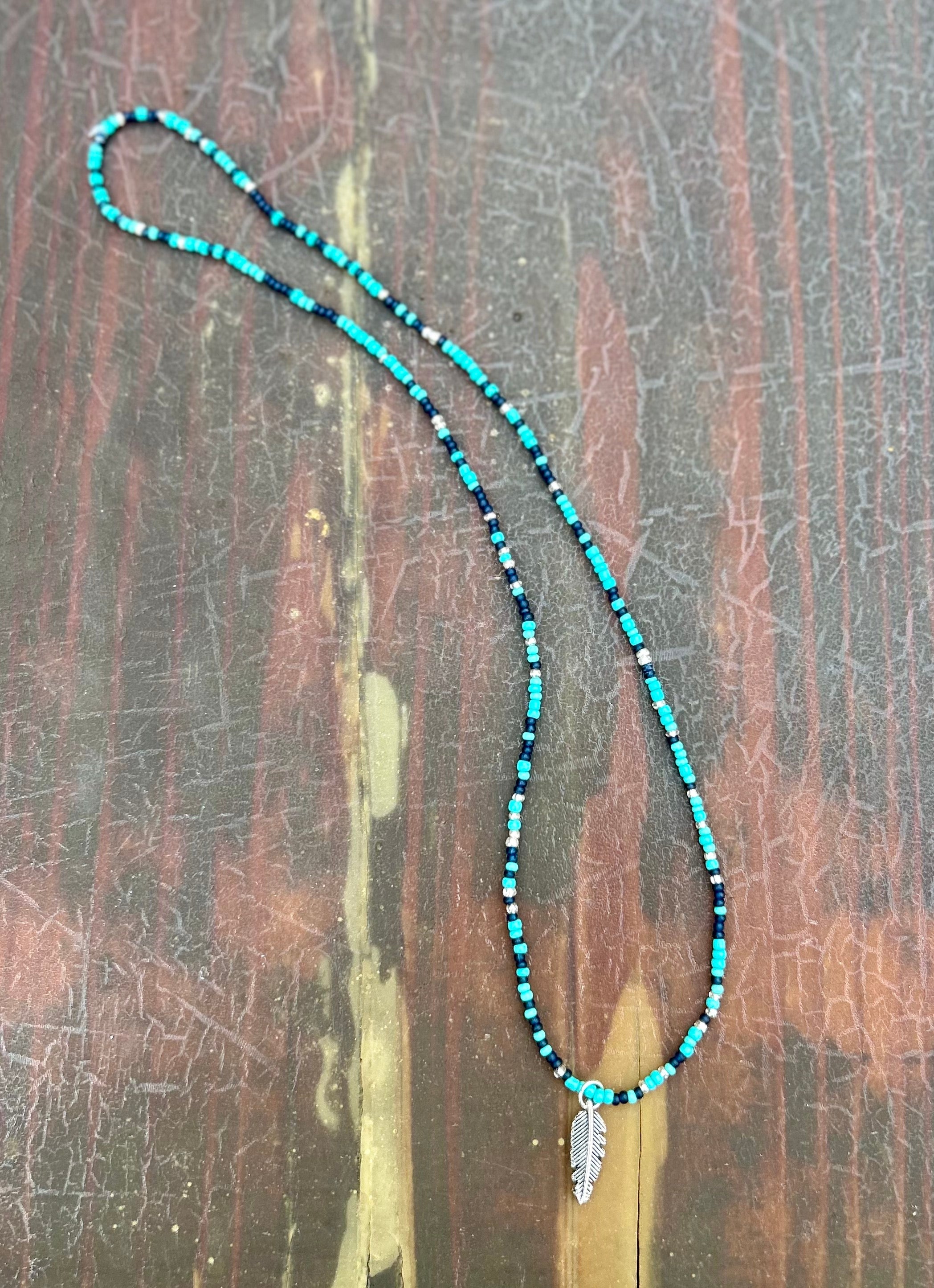 A vibrant Mixed Turquoise Navy Boho Seed Bead Necklace featuring a silver feather pendant, showcasing colorful Czech glass seed beads on elastic cording.