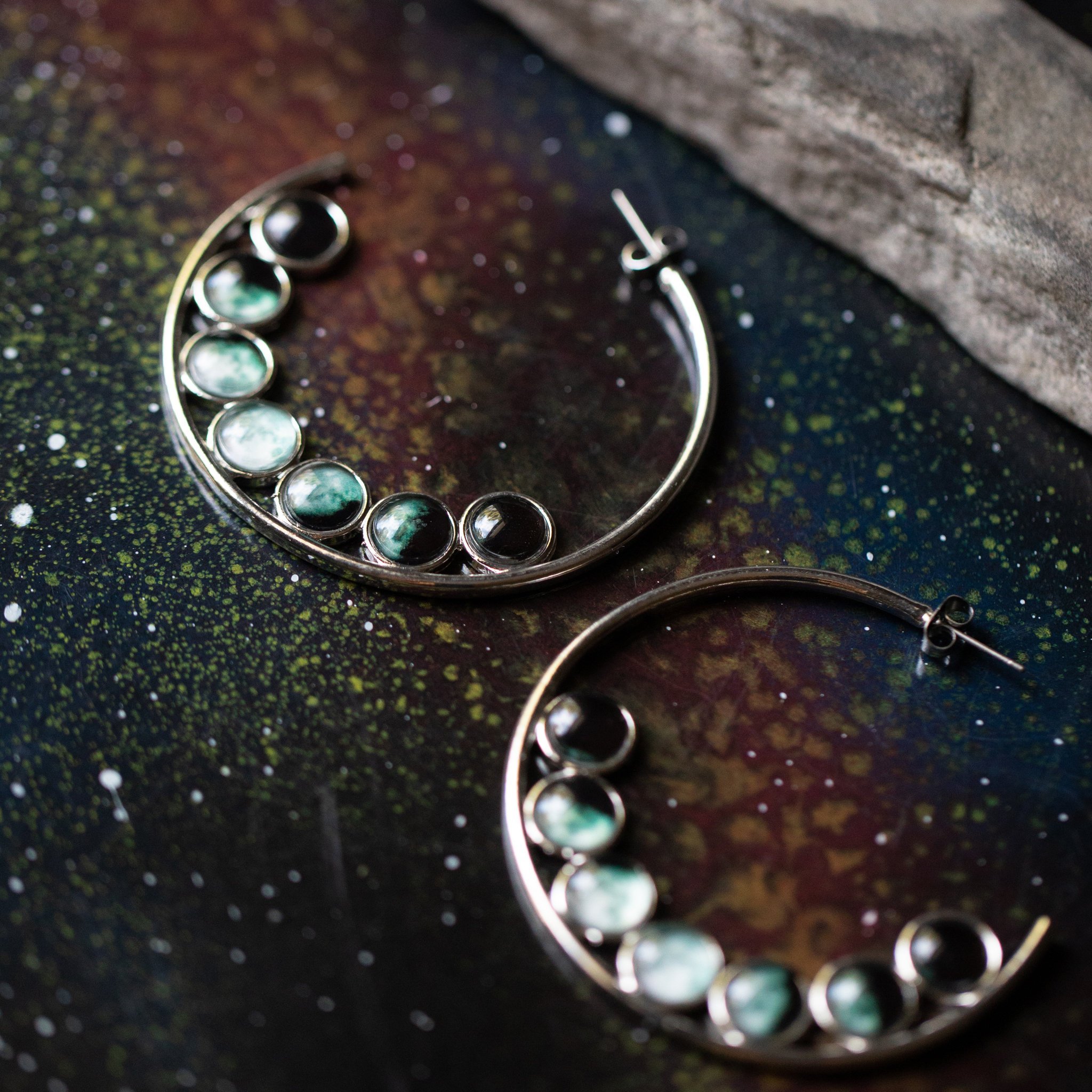 A pair of silver-tone hoop earrings featuring seven phases of the Moon, handcrafted with a textured metal back.