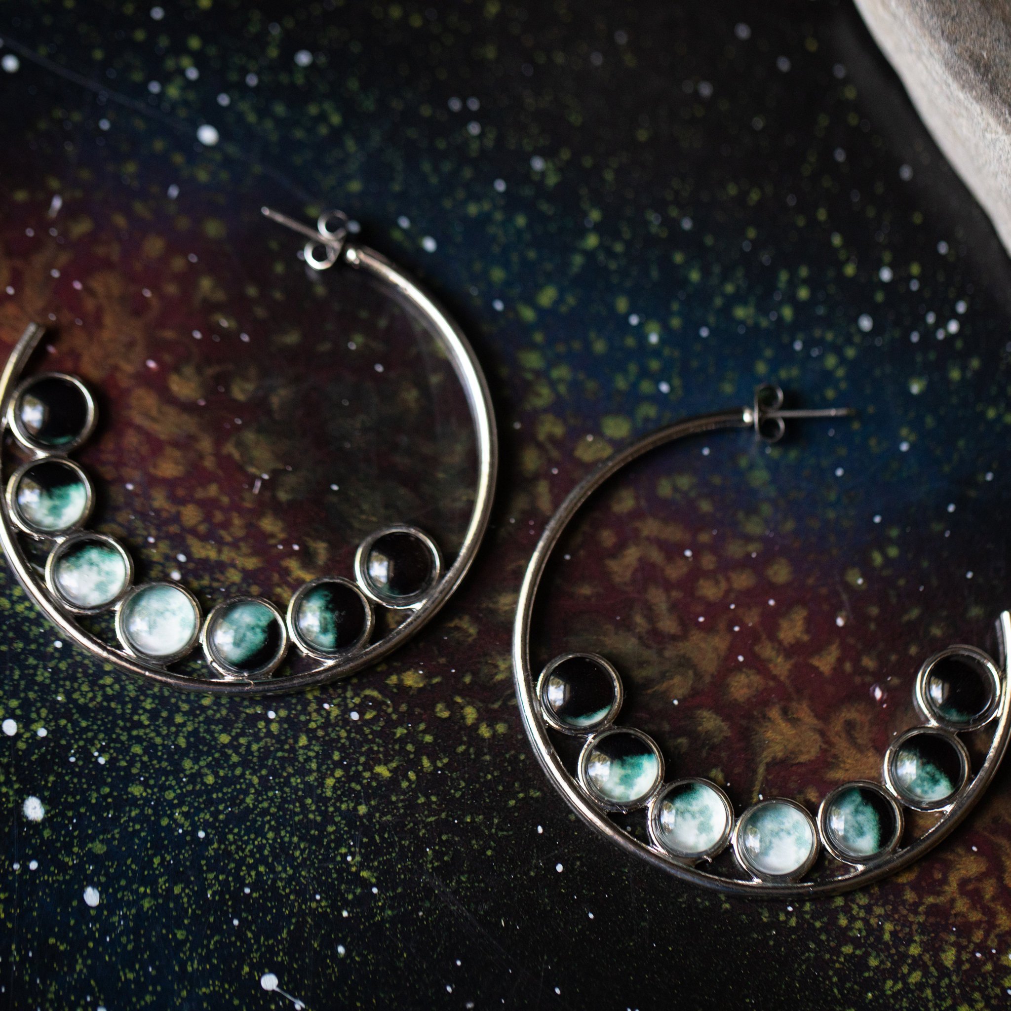 A pair of silver-tone hoop earrings featuring seven phases of the Moon, handcrafted with a textured metal back.