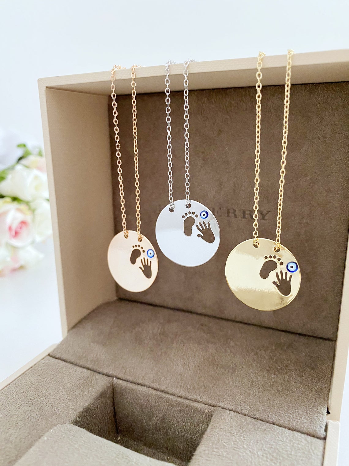 New Baby Necklace featuring an Evil Eye design with baby feet and handprint motifs, available in gold, silver, and rose gold.