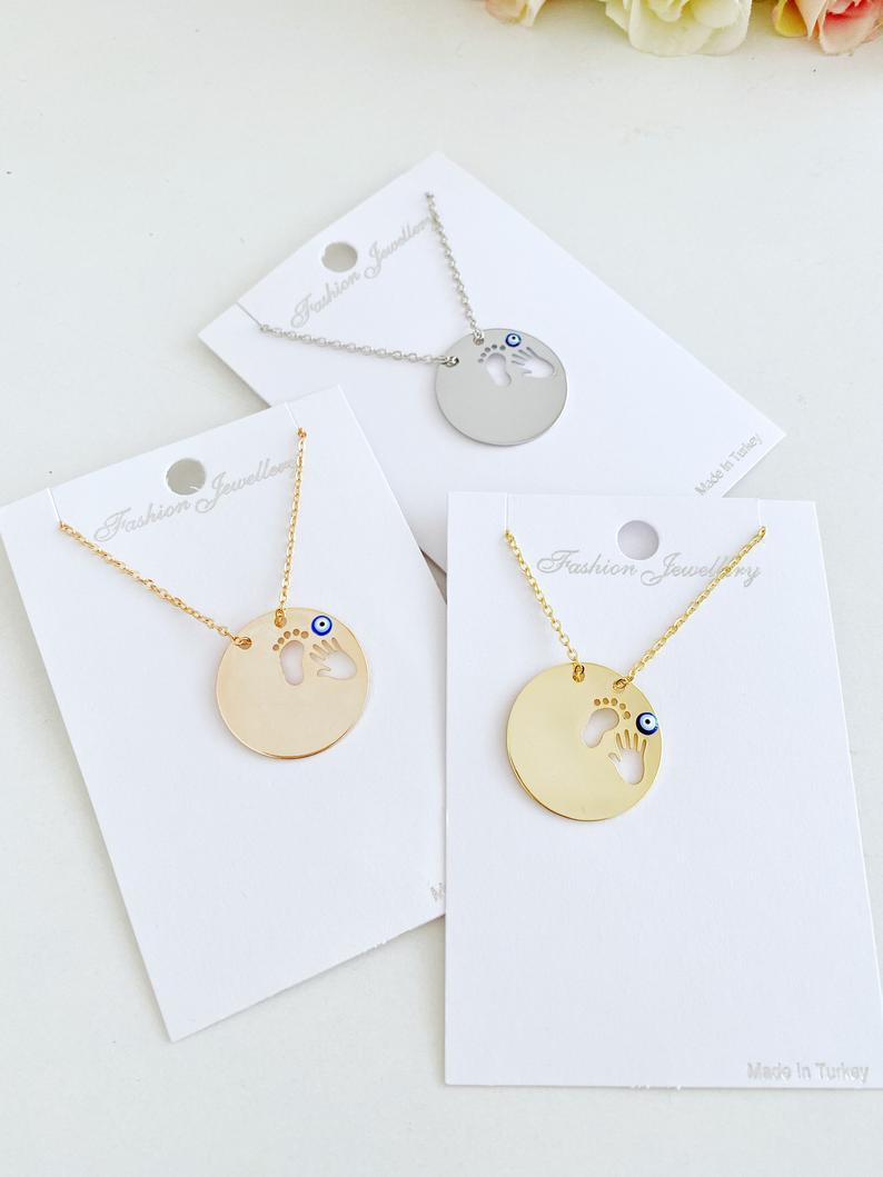 New Baby Necklace featuring an Evil Eye design with baby feet and handprint motifs, available in gold, silver, and rose gold.