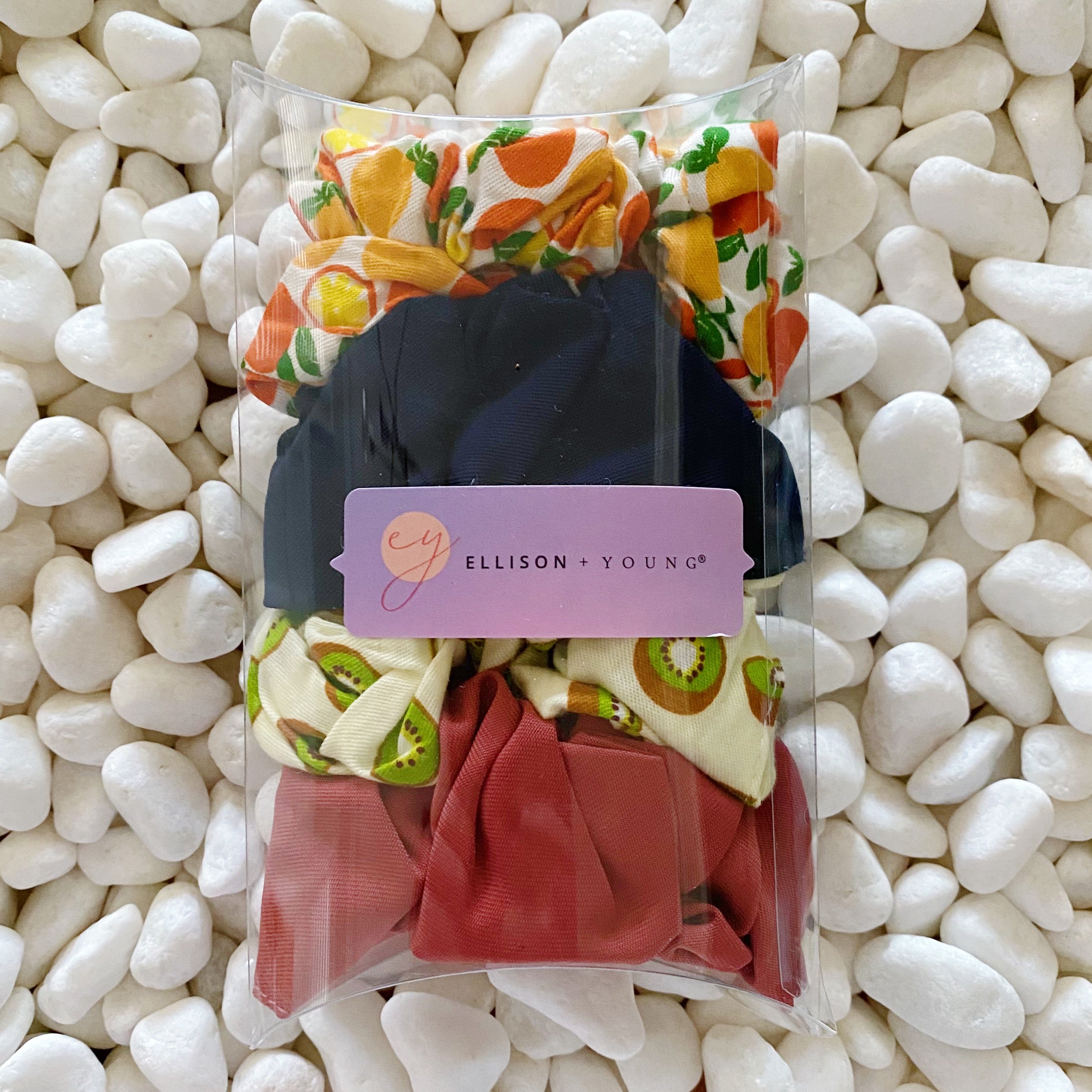 A set of four colorful hair scrunchies featuring two fruit prints and two solid colors, neatly arranged in a clear cello case.