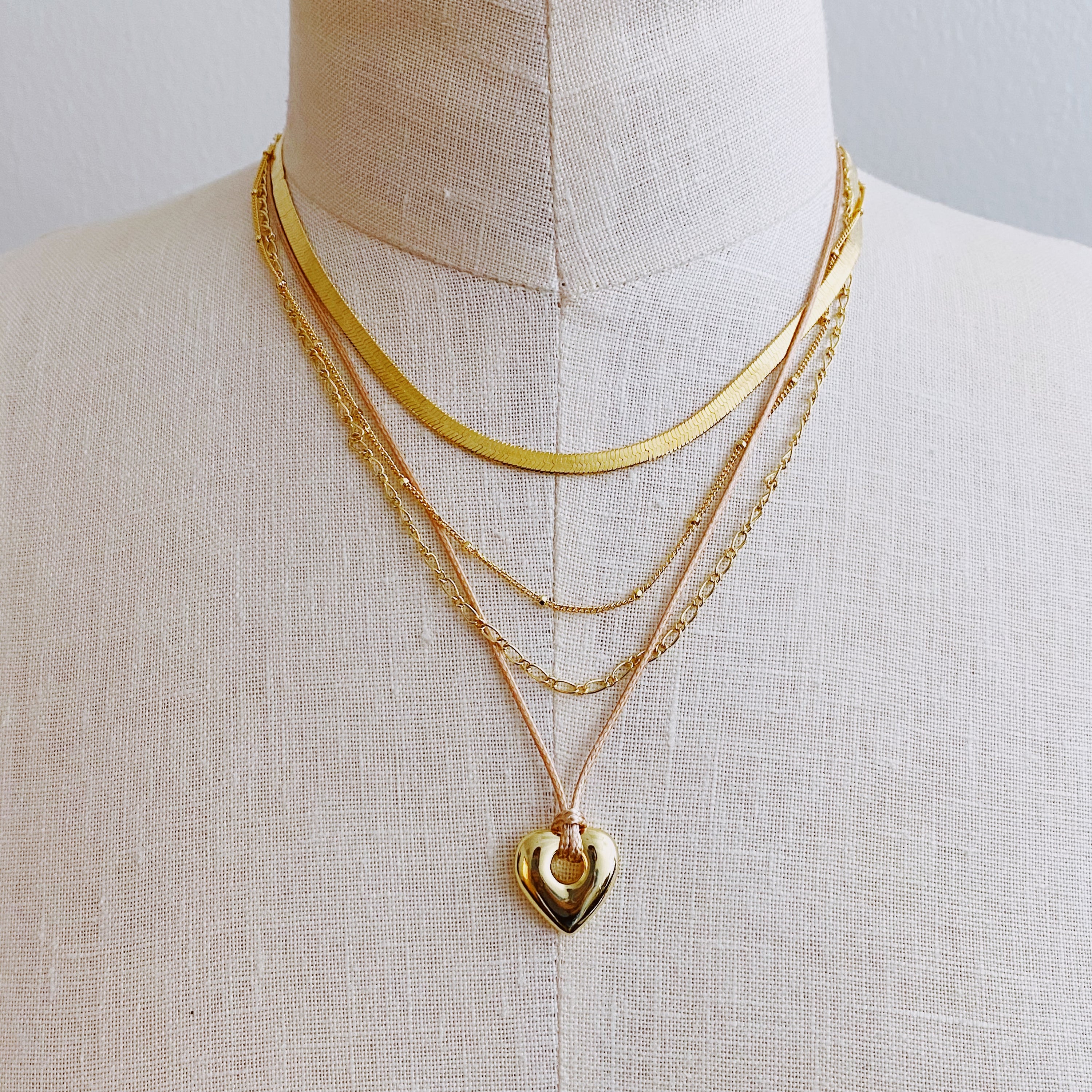 A beautifully layered heart and chain necklace featuring four strands, with a heart pendant and dainty metal chains in 18K gold plating.