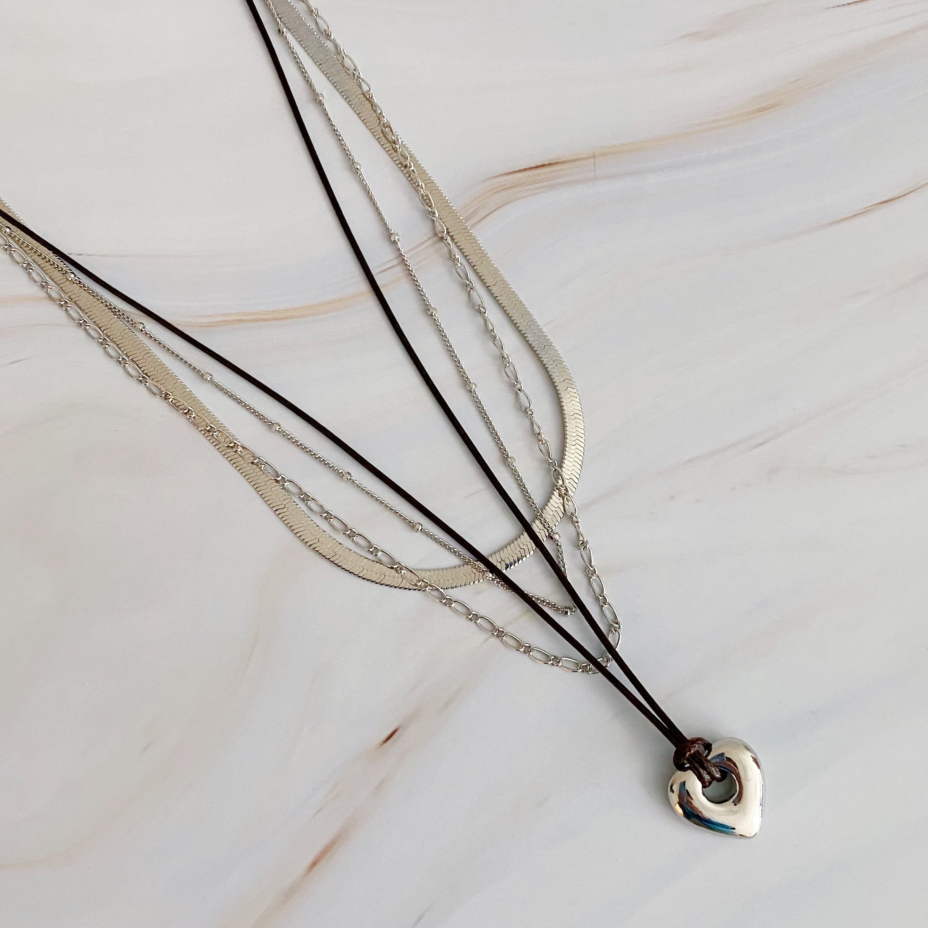 A beautifully layered heart and chain necklace featuring four strands, with a heart pendant and dainty metal chains in 18K gold plating.