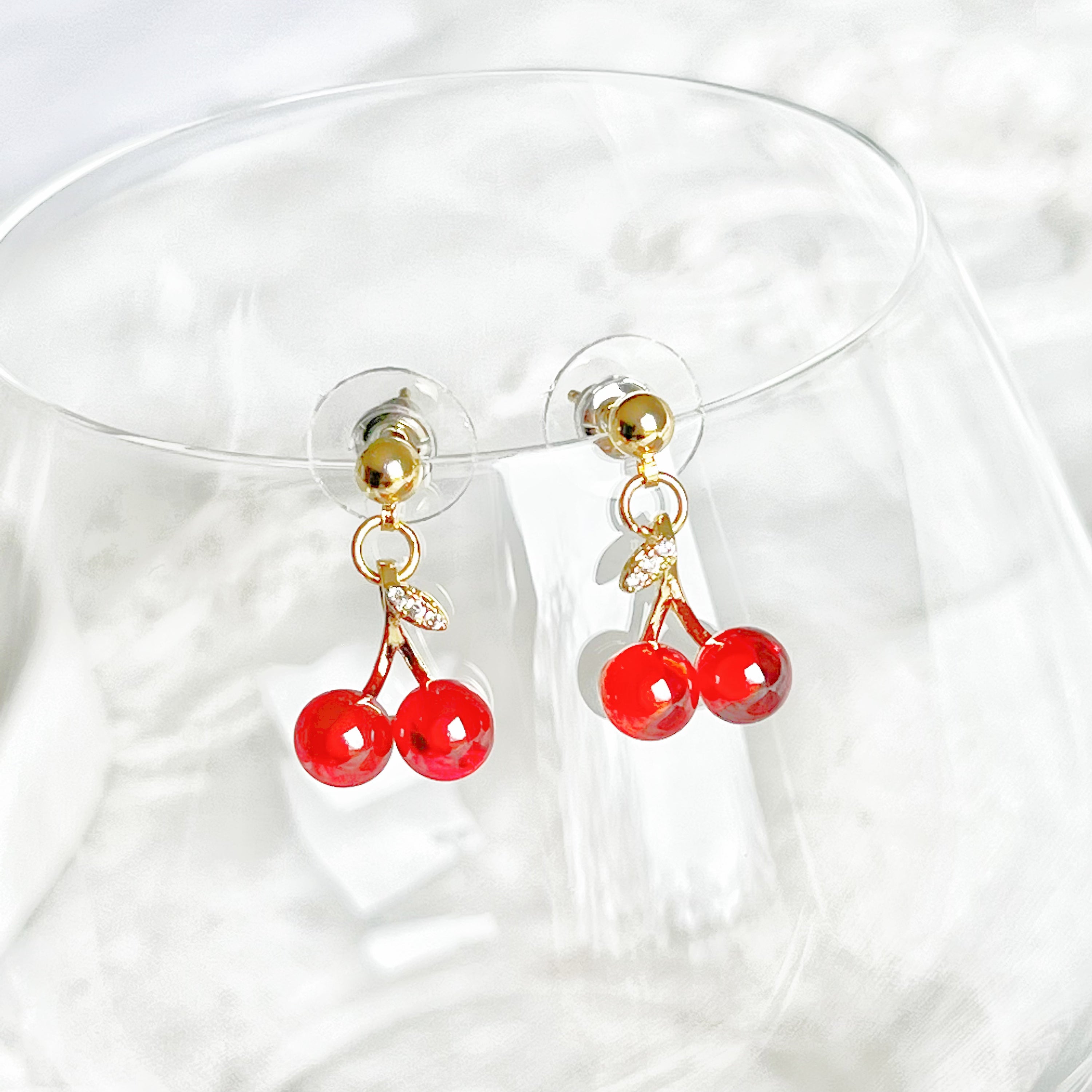 A pair of elegant Red Cherry Mini Drop Earrings featuring red zircon cherries and clear zircon bow ties, set on a gold-plated bronze base.