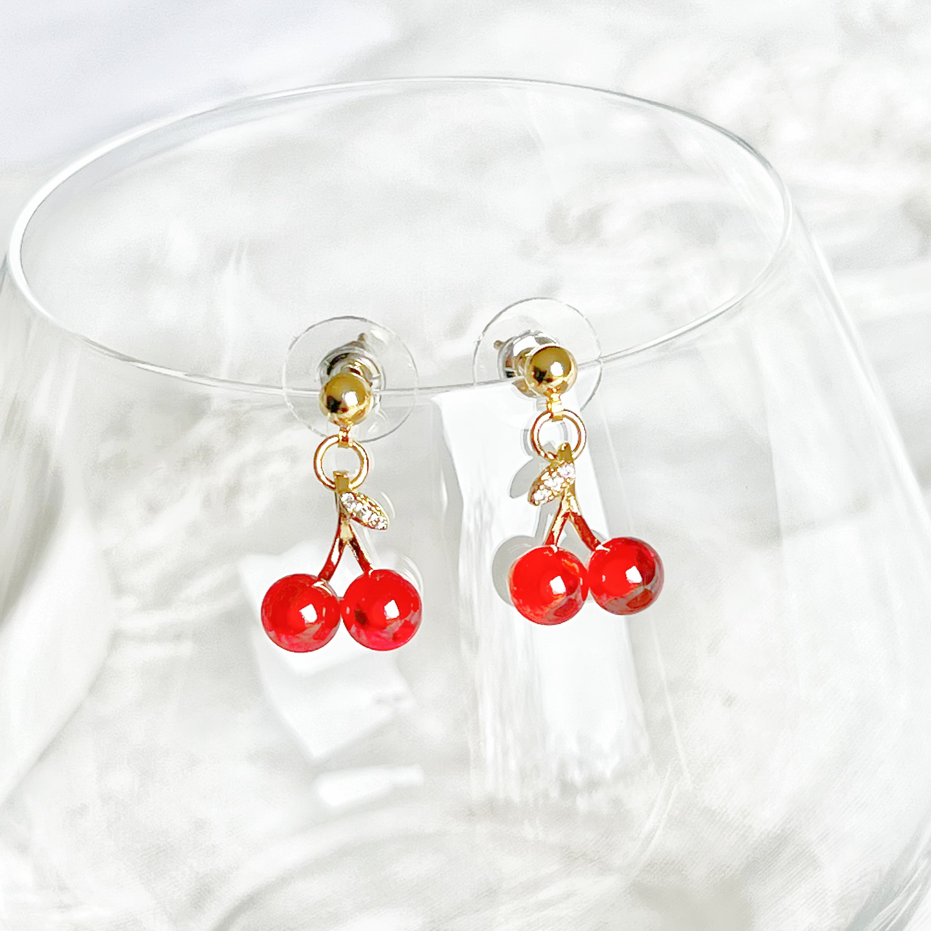 A pair of elegant Red Cherry Mini Drop Earrings featuring red zircon cherries and clear zircon bow ties, set on a gold-plated bronze base.