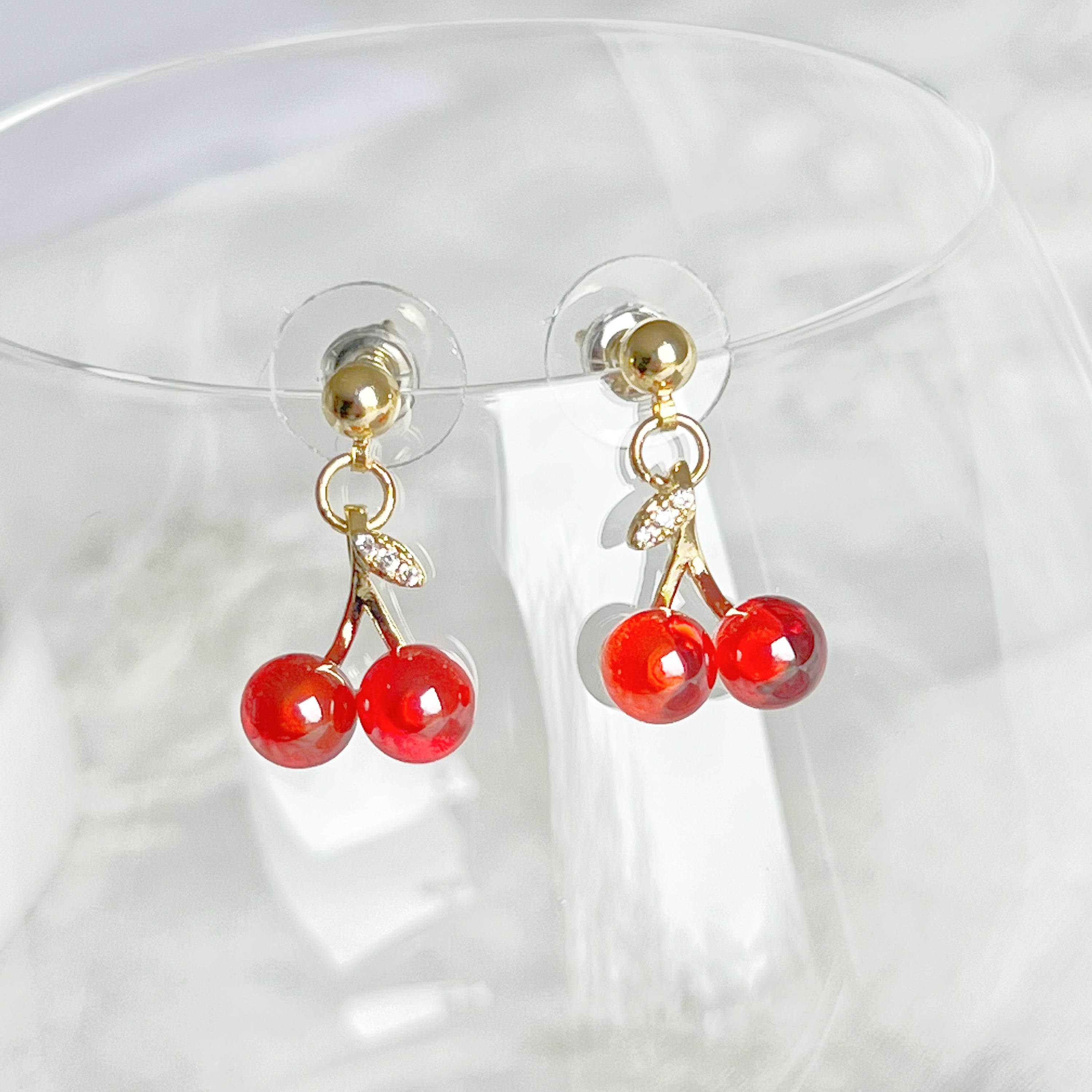 A pair of elegant Red Cherry Mini Drop Earrings featuring red zircon cherries and clear zircon bow ties, set on a gold-plated bronze base.