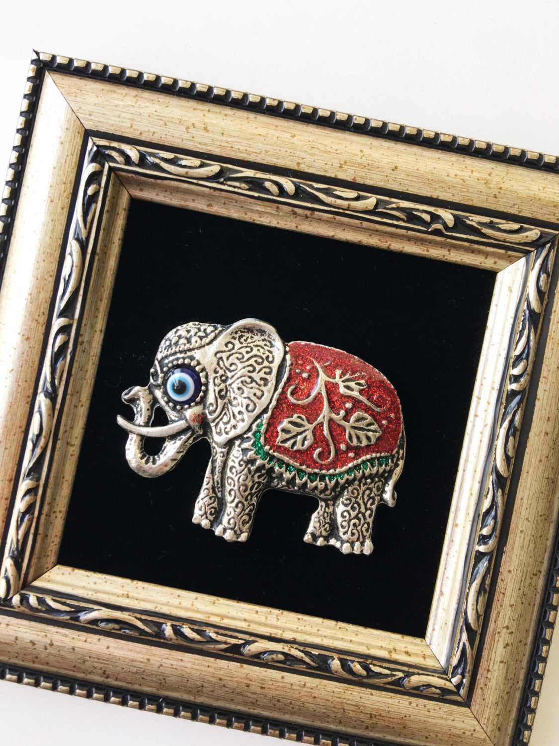 A handmade Red Elephant Frame Wall Hanging featuring a silver elephant and a small evil eye bead, measuring 14cm x 14cm.