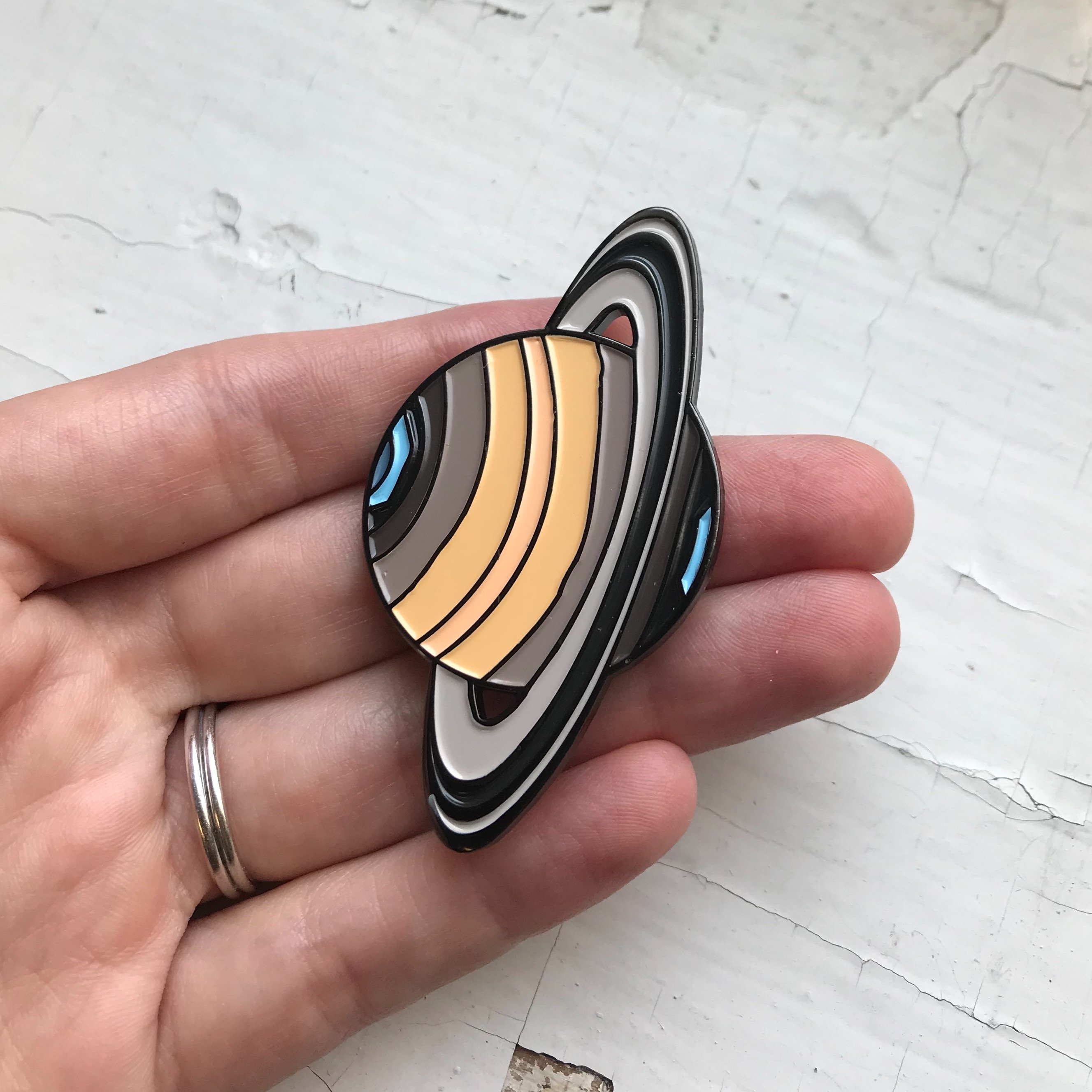 A colorful Saturn enamel pin featuring the planet with rings, designed by Lauren Beacham, perfect for jackets and backpacks.