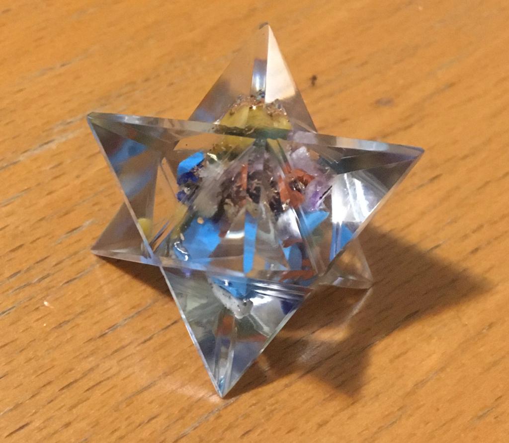 A Seven Chakra Orgone Merkaba made of translucent resin with seven colorful chakra gemstone chips, shaped like an eight-pointed star.