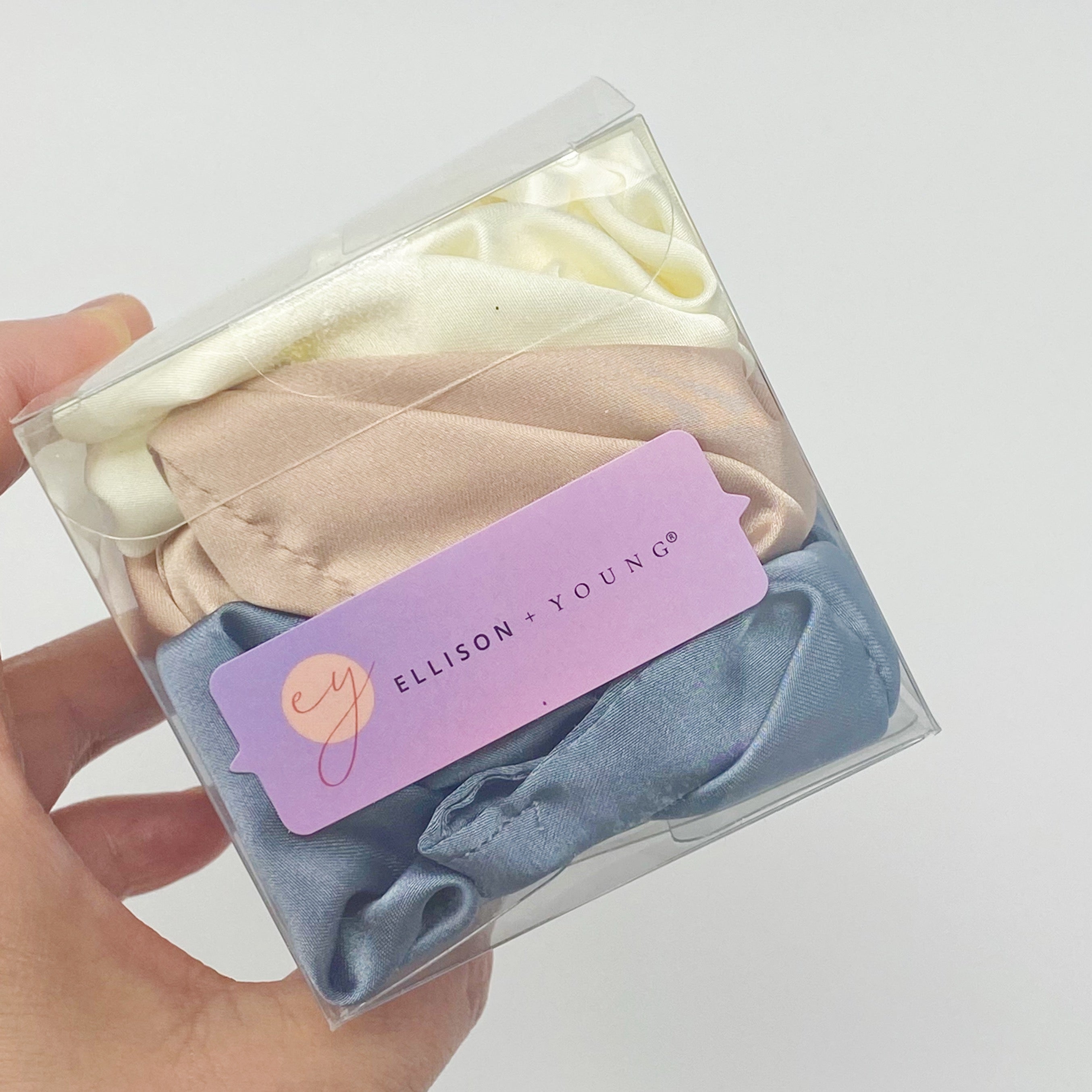 Set of 3 soft satin scrunchies in various colors, elegantly packaged in a clear cello case with Ellison + Young branding.