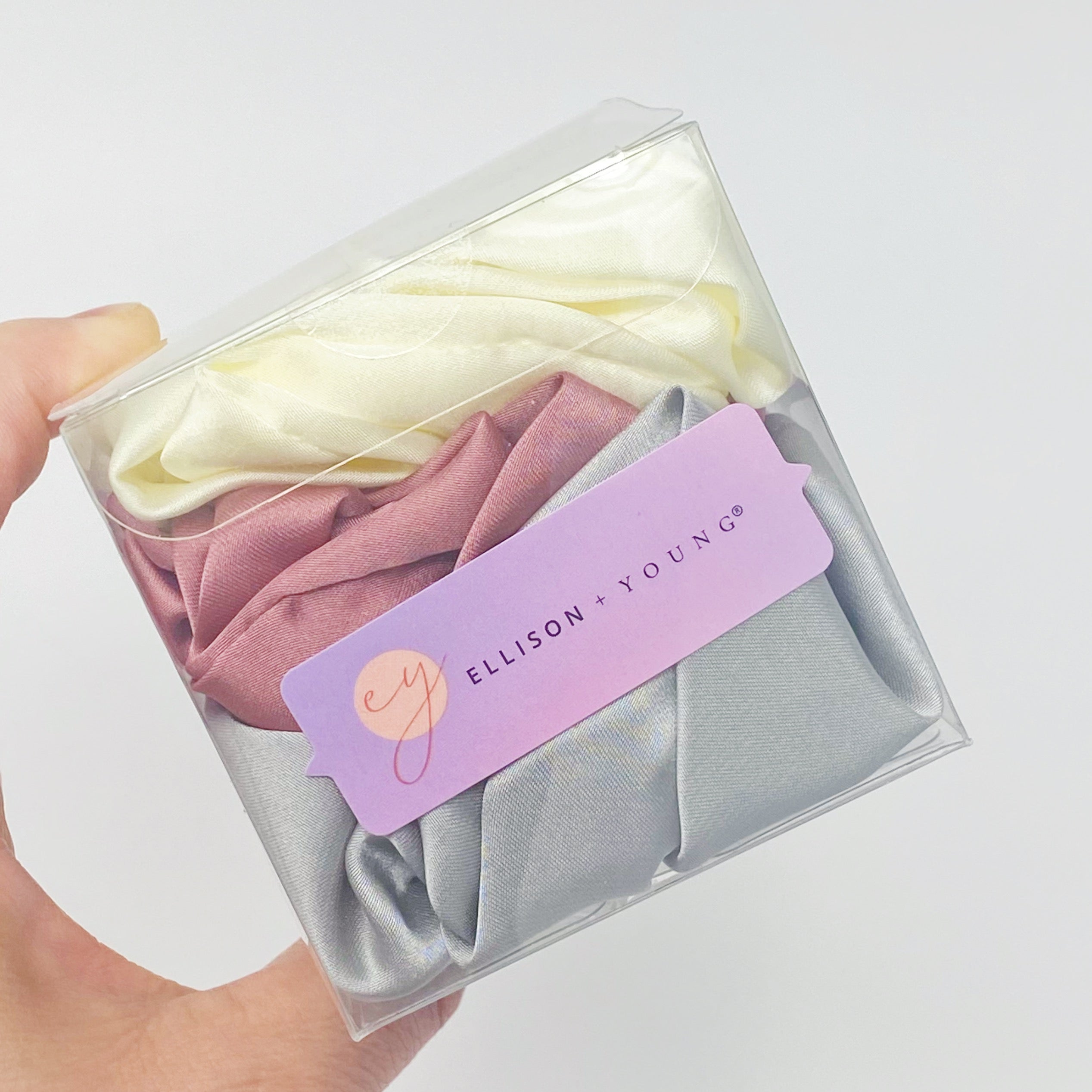 Set of 3 soft satin scrunchies in various colors, elegantly packaged in a clear cello case with Ellison + Young branding.