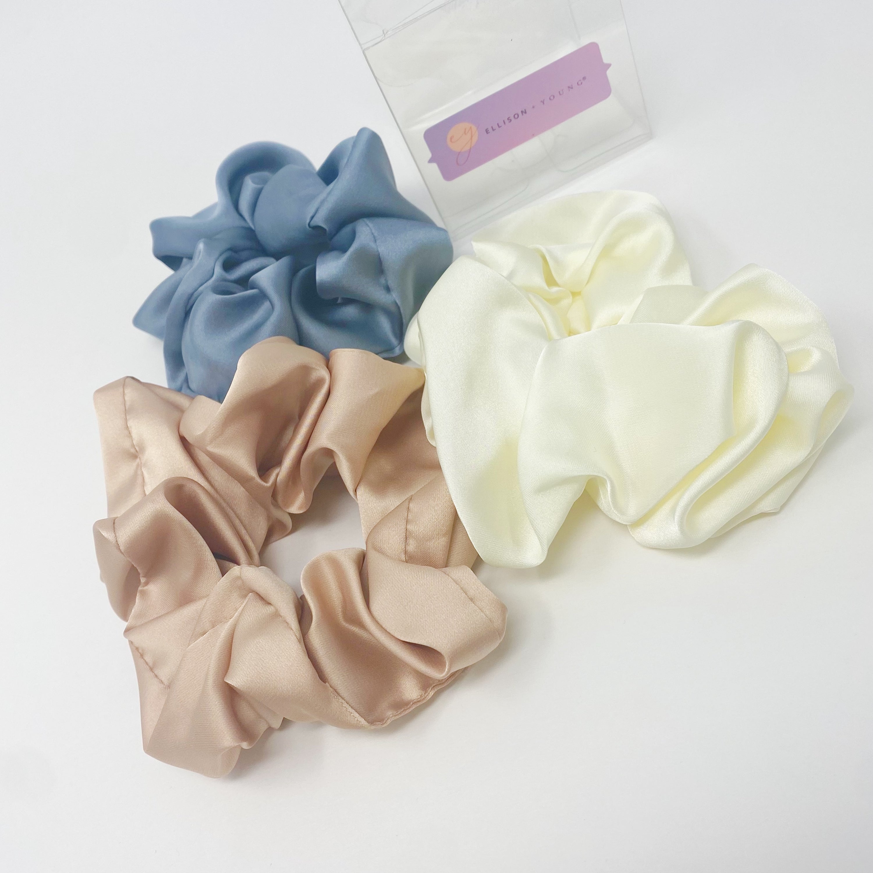 Set of 3 soft satin scrunchies in various colors, elegantly packaged in a clear cello case with Ellison + Young branding.