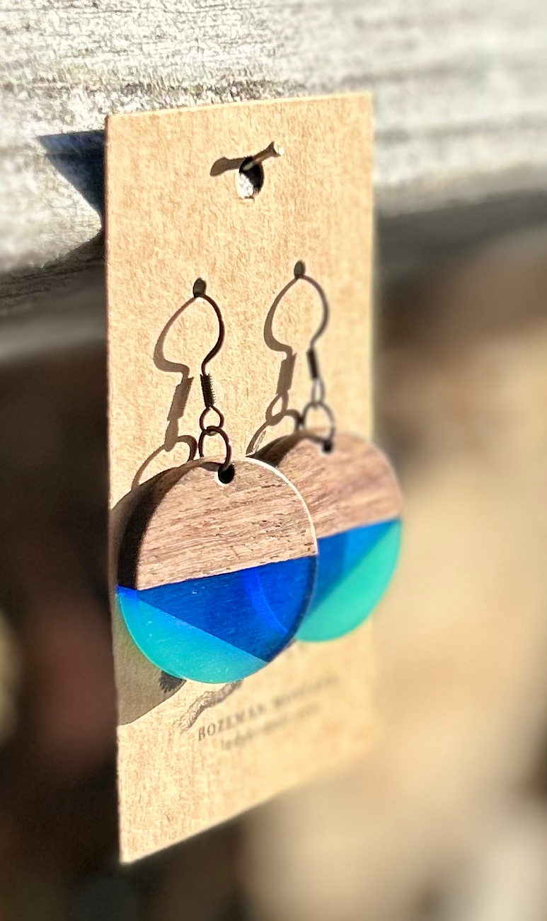 A pair of circle earrings made from walnut wood and blue resin, showcasing a geometric pattern and natural wood grain.