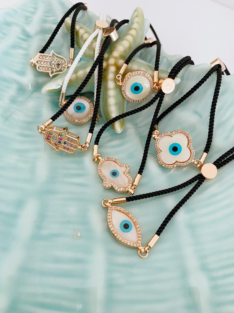 A stylish White Evil Eye Bracelet featuring a Hamsa charm on a black string, adorned with zirconia around a clover charm.