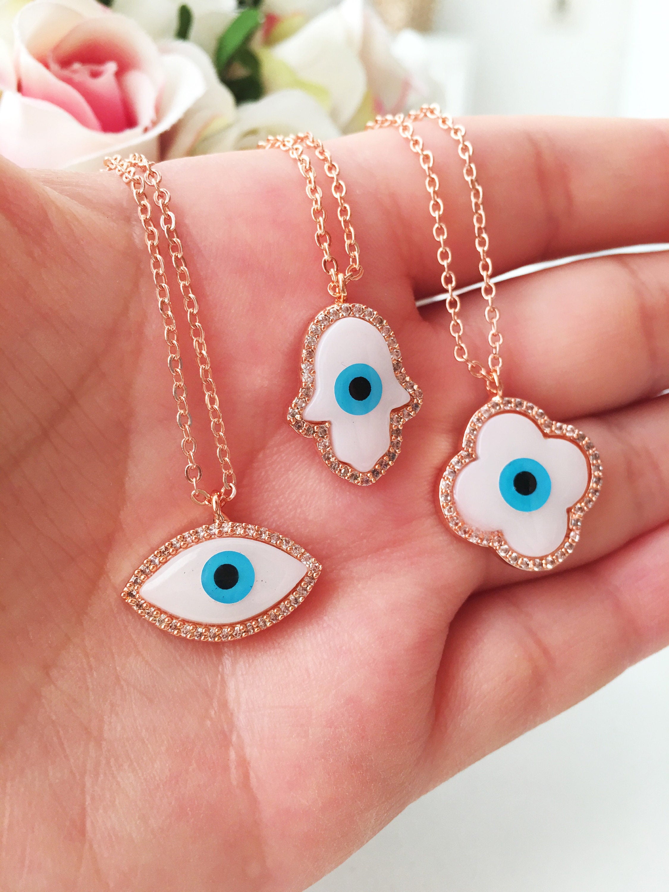 A beautiful White Evil Eye Necklace featuring a Hamsa Hand and Clover Charm, showcasing intricate details and a rose gold finish.