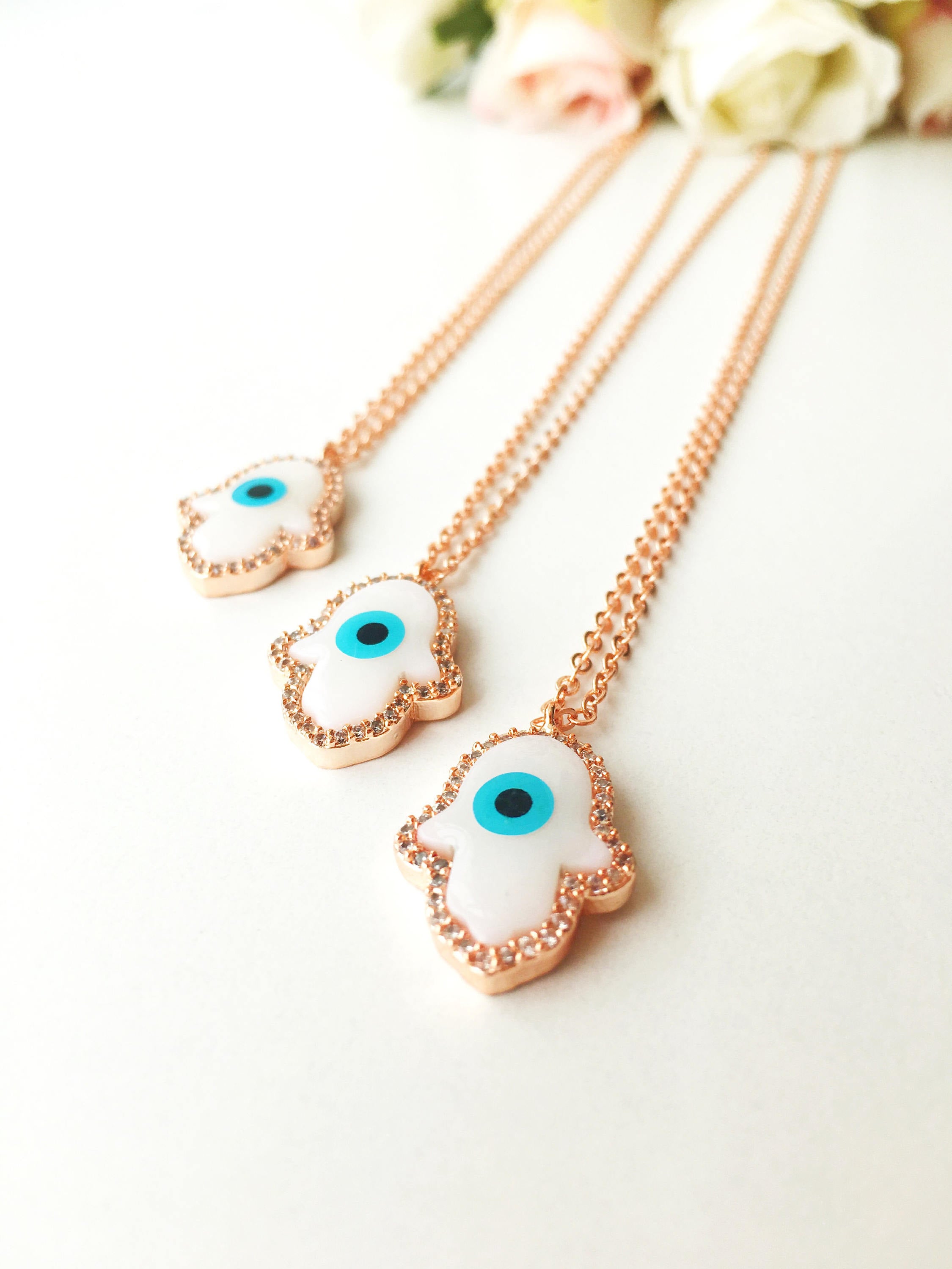 A beautiful White Evil Eye Necklace featuring a Hamsa Hand and Clover Charm, showcasing intricate details and a rose gold finish.