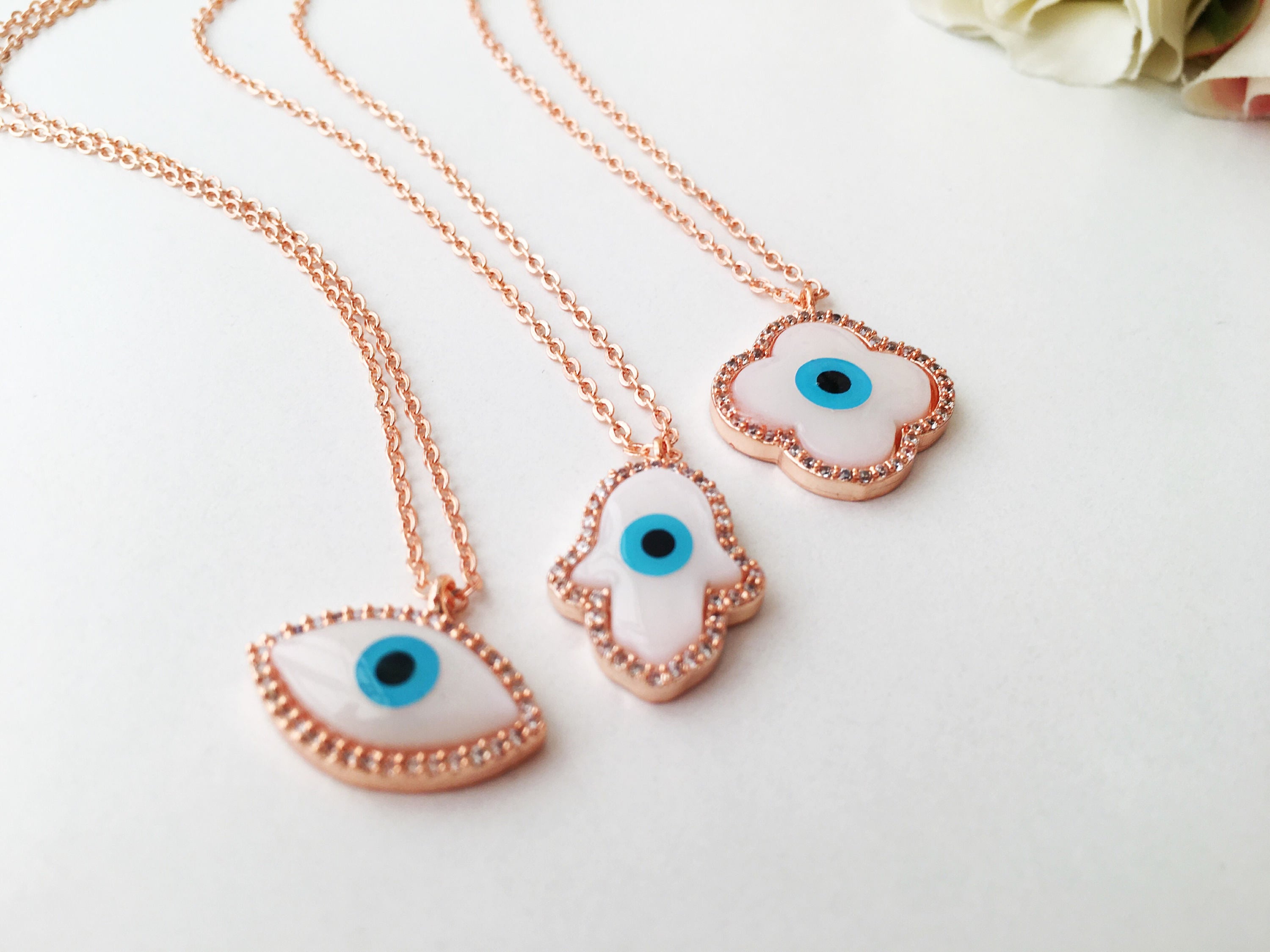 A beautiful White Evil Eye Necklace featuring a Hamsa Hand and Clover Charm, showcasing intricate details and a rose gold finish.