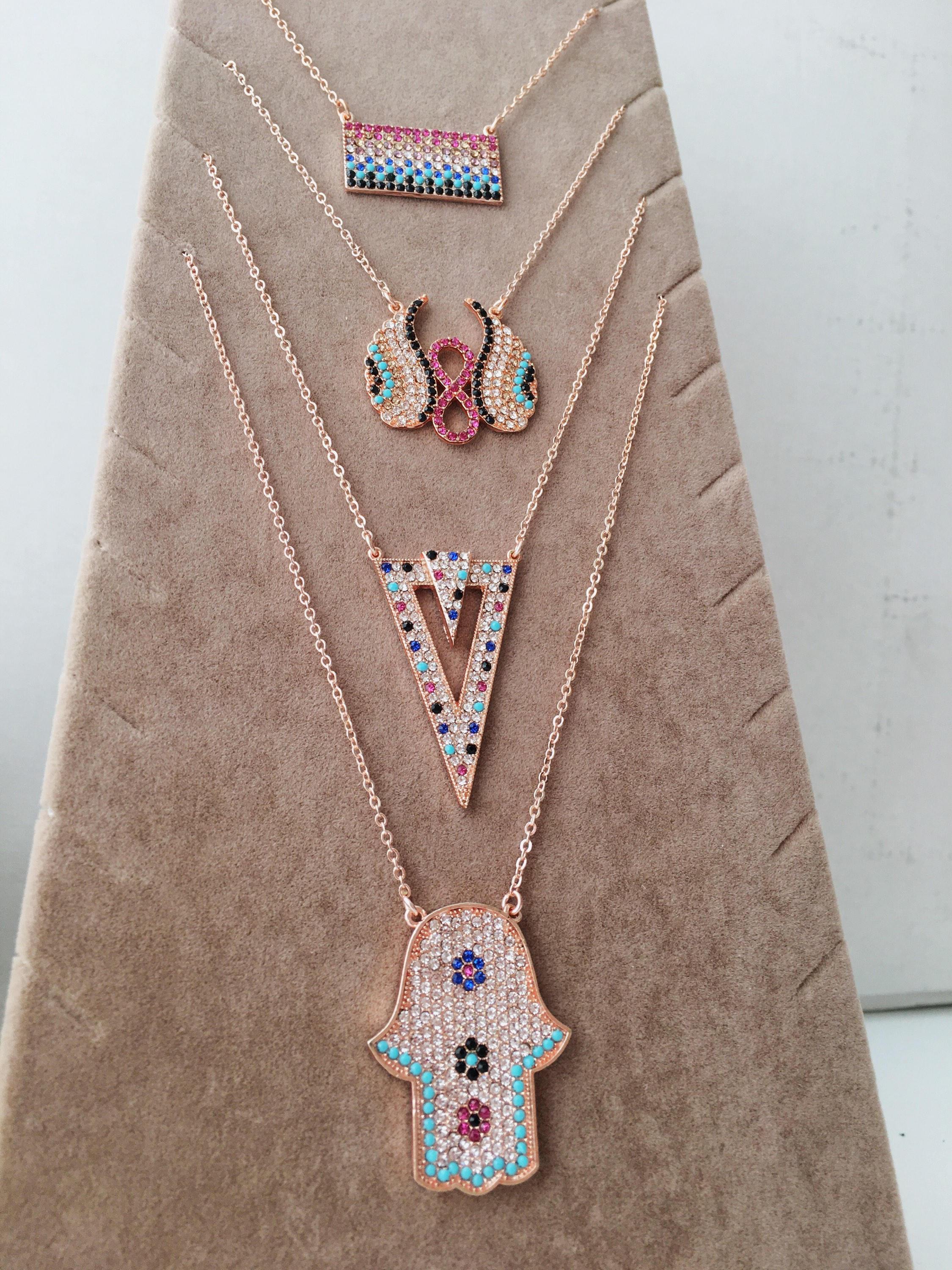 A collection of handmade zirconia necklaces featuring hamsa hand, angel wings, and rectangular designs, crafted in rose gold stainless steel.