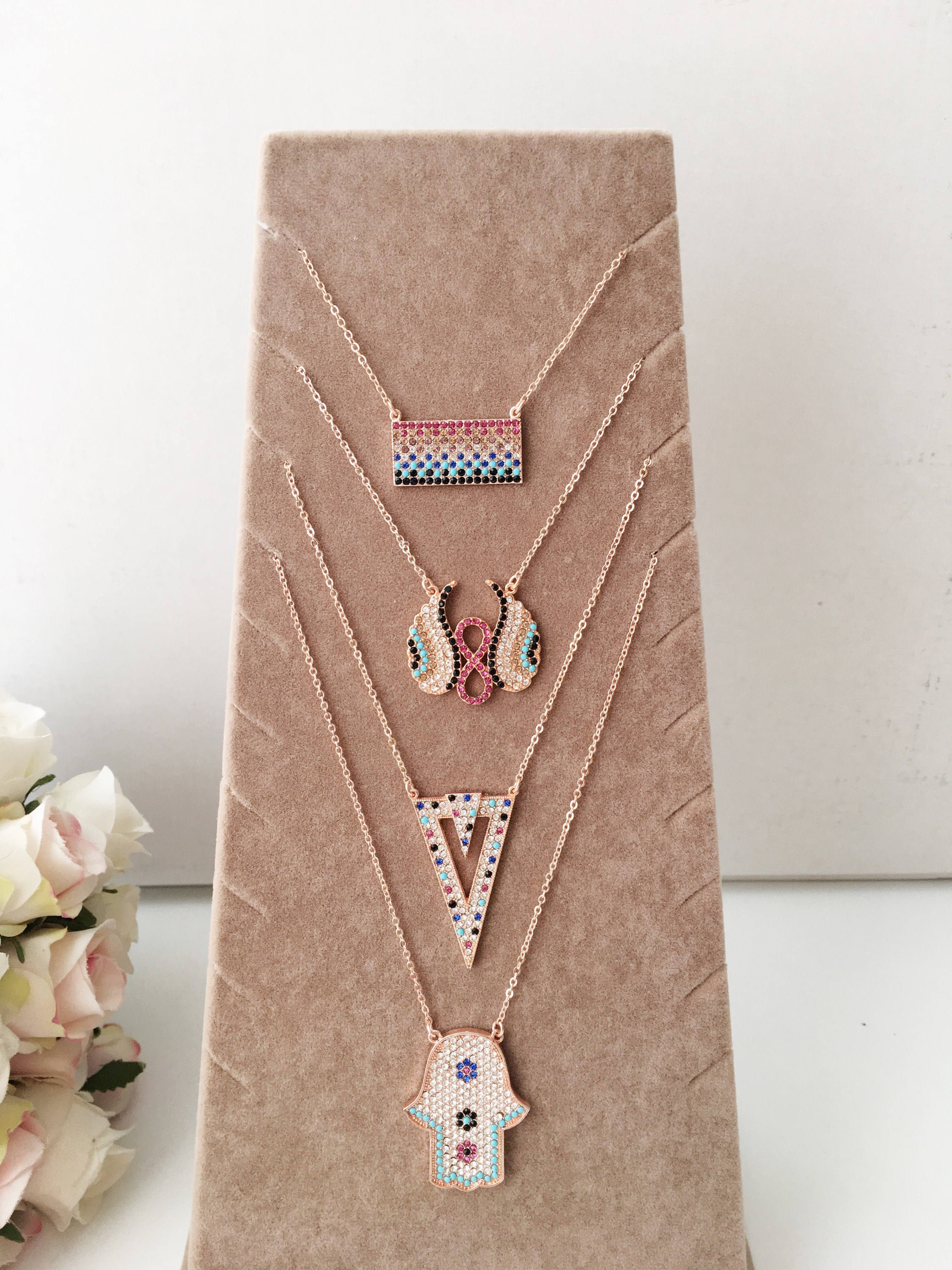 A collection of handmade zirconia necklaces featuring hamsa hand, angel wings, and rectangular designs, crafted in rose gold stainless steel.