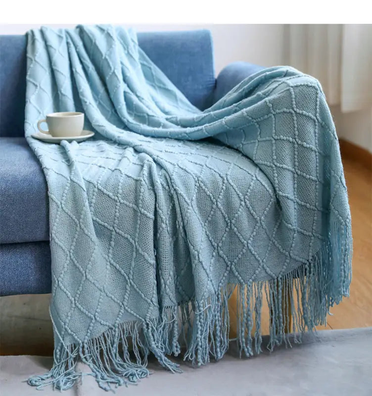 A cozy lake blue knitted throw blanket with tassels, featuring a classic diamond pattern, elegantly draped over a sofa.