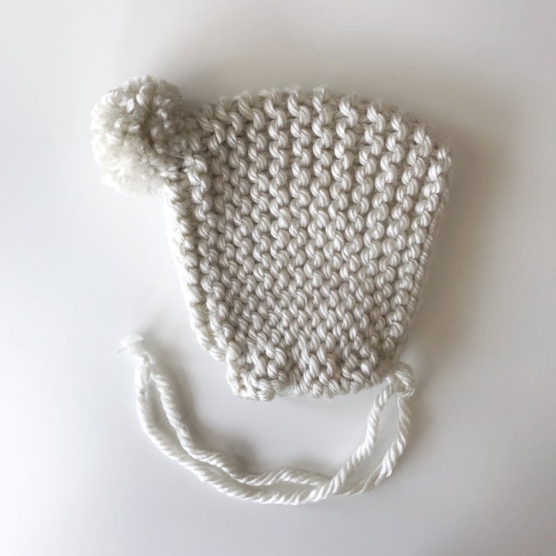 A cream-colored handmade knit bonnet with a pom on top, designed to stretch and fit comfortably on any head shape.