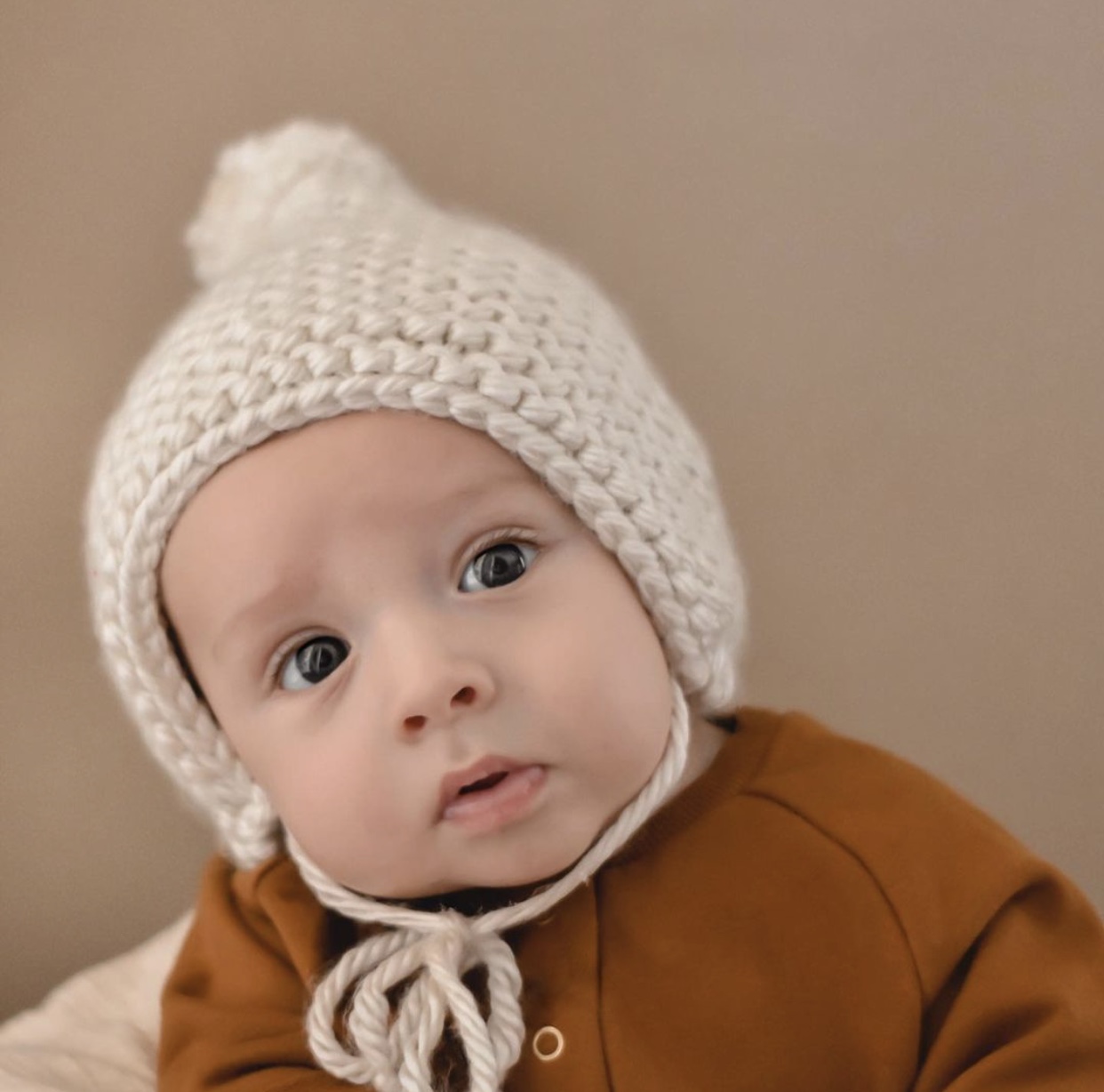 A cream-colored handmade knit bonnet with a pom on top, designed to stretch and fit comfortably on any head shape.