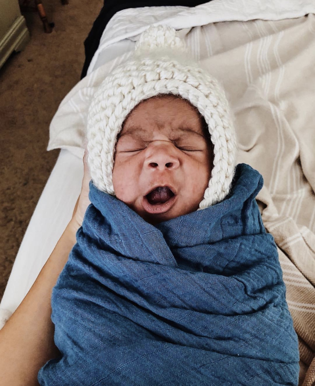 A cream-colored handmade knit bonnet with a pom on top, designed to stretch and fit comfortably on any head shape.