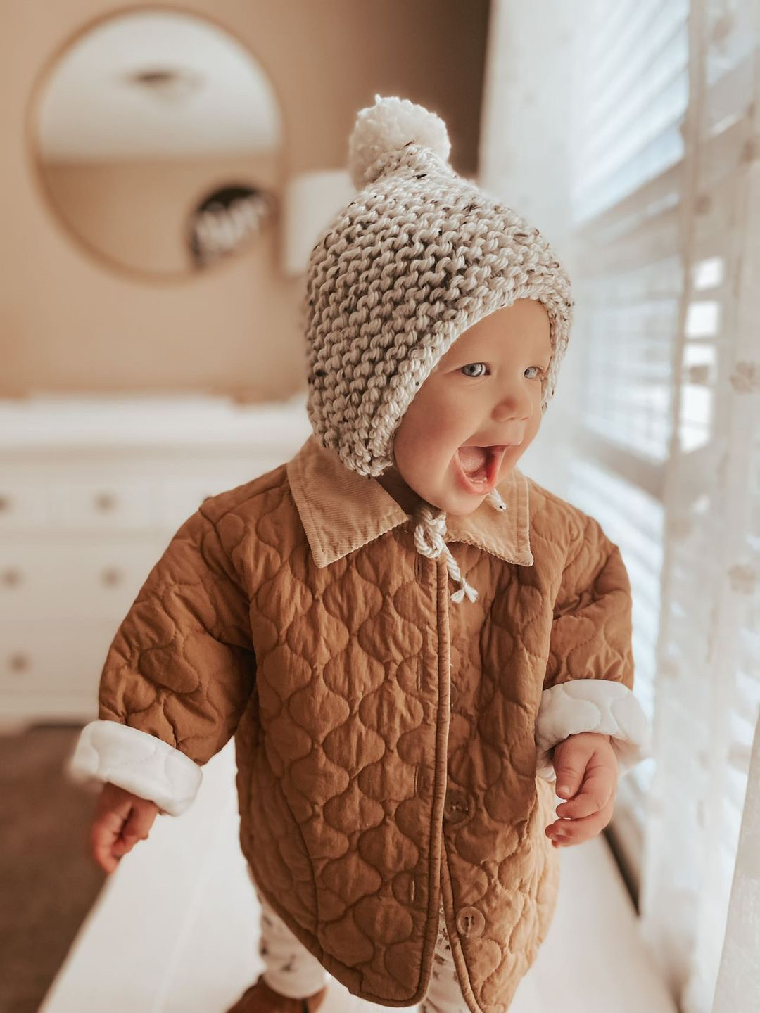 A cozy oatmeal temperance bonnet with a pom detail, showcasing its handmade knit design and stretchy fit.